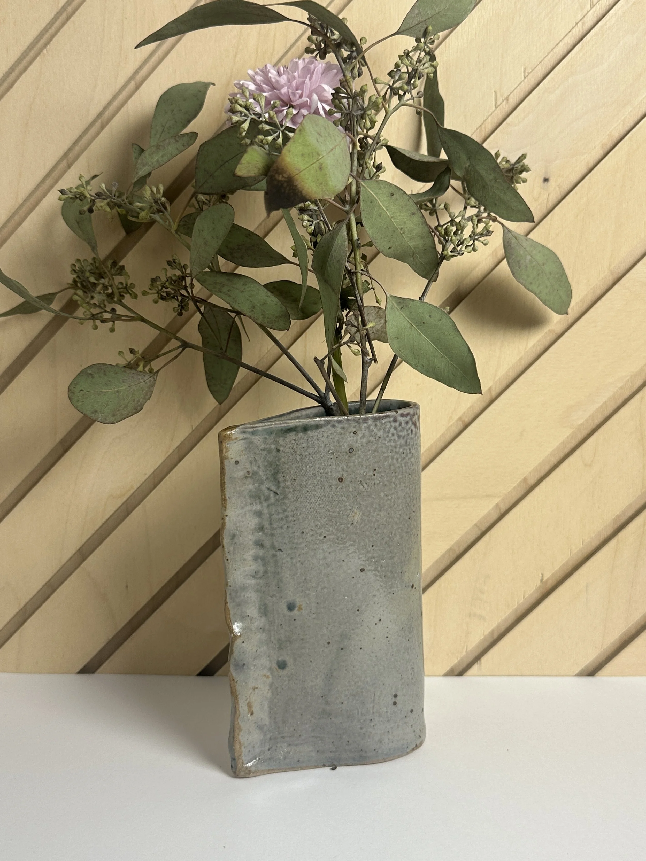 A ceramic vase with a grayish glaze holding a bouquet of green leaves, small flowers, and a single pink flower, placed on a white surface against a light wooden wall background.