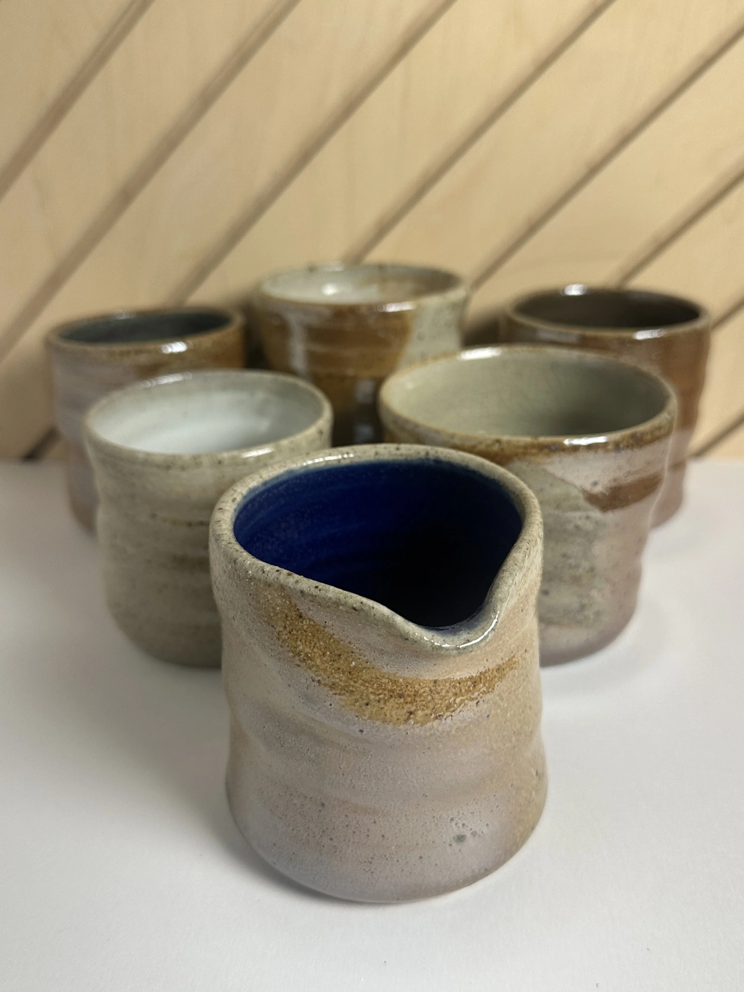 Set of handmade ceramic cups with various glaze colors, arranged on a white surface with a wooden background. These are all completely unique and soda fired.