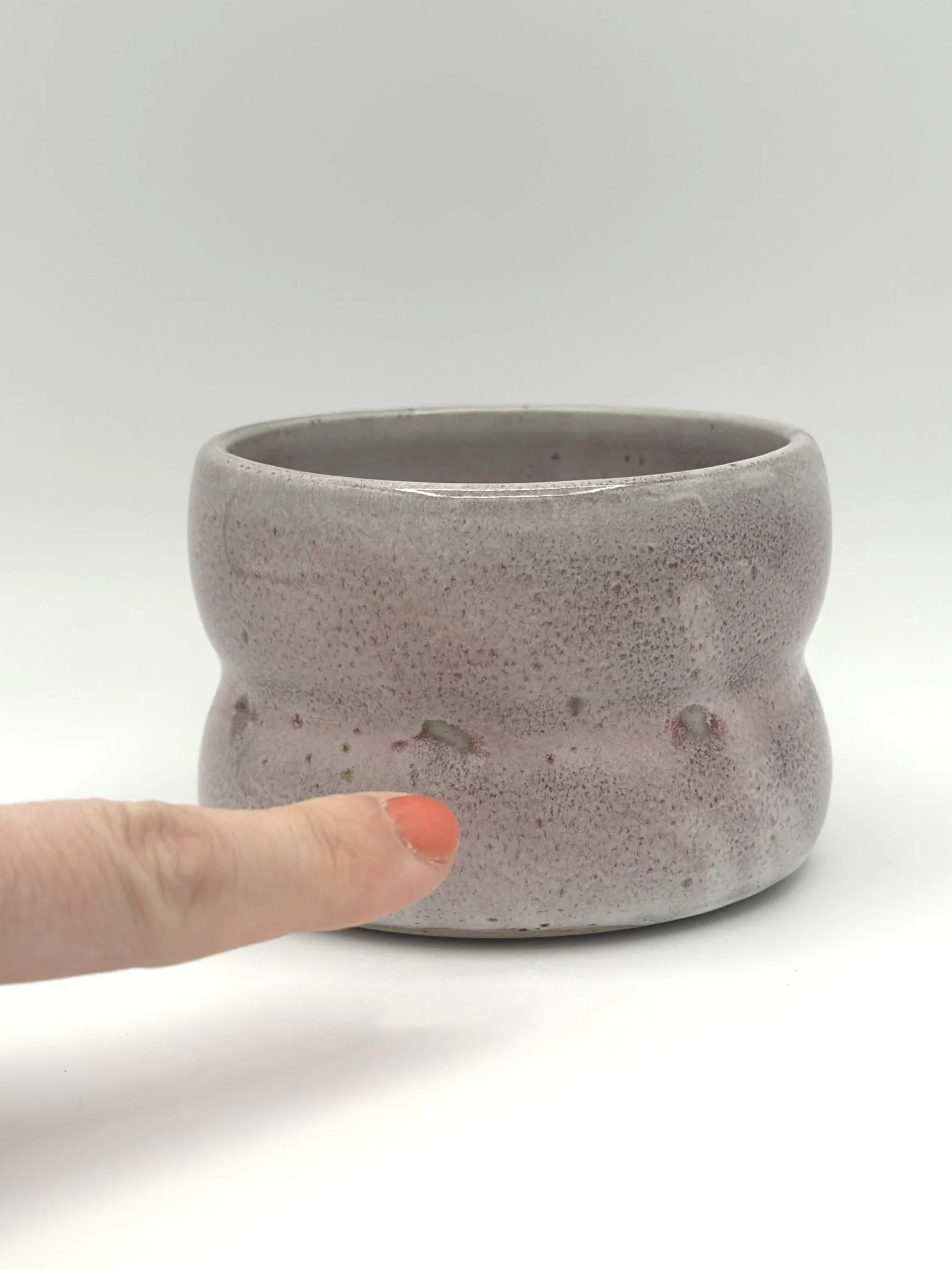 Handmade stoneware ceramic tumbler with 2 bubbles and a light grayish and pinkish candy coated shiny glaze. manicured finger pointing to glaze imperfections