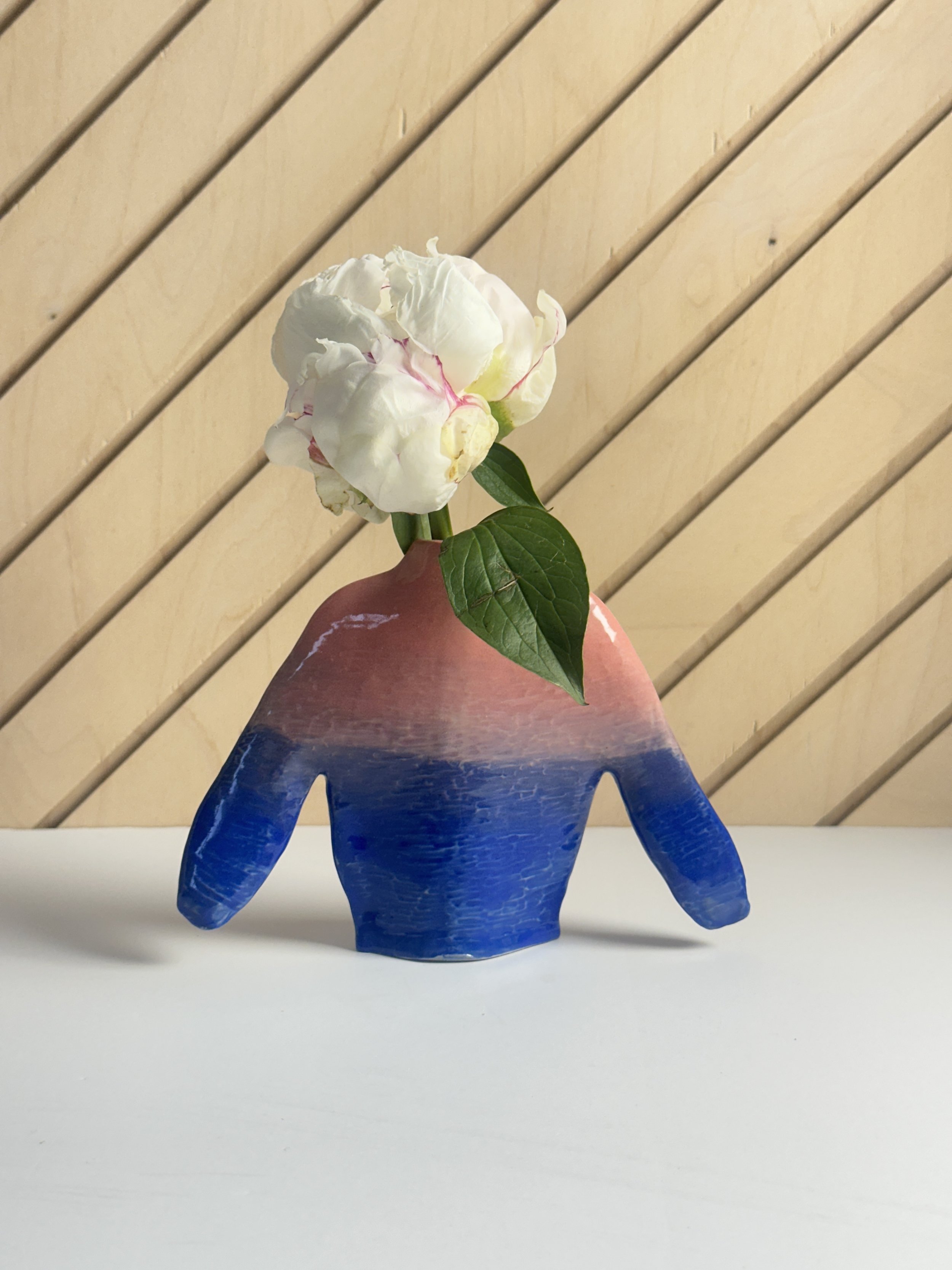 A colorful glass vase shaped like a hand with a green leaf and white flower inside, placed on a white surface with a wooden slat wall background.