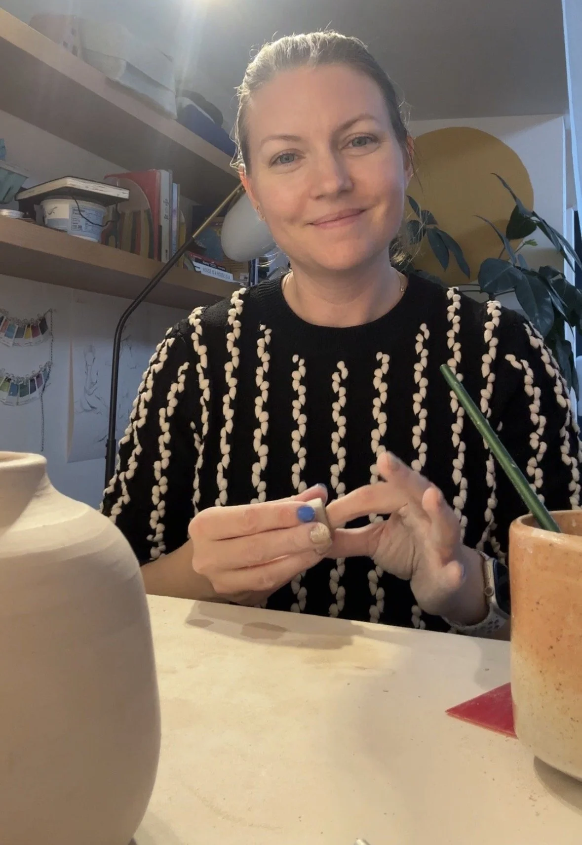 A woman with light skin and blonde hair tied back, smiling while sitting at a table. She is wearing a black top with white swirling patterns and has clay in her hands.