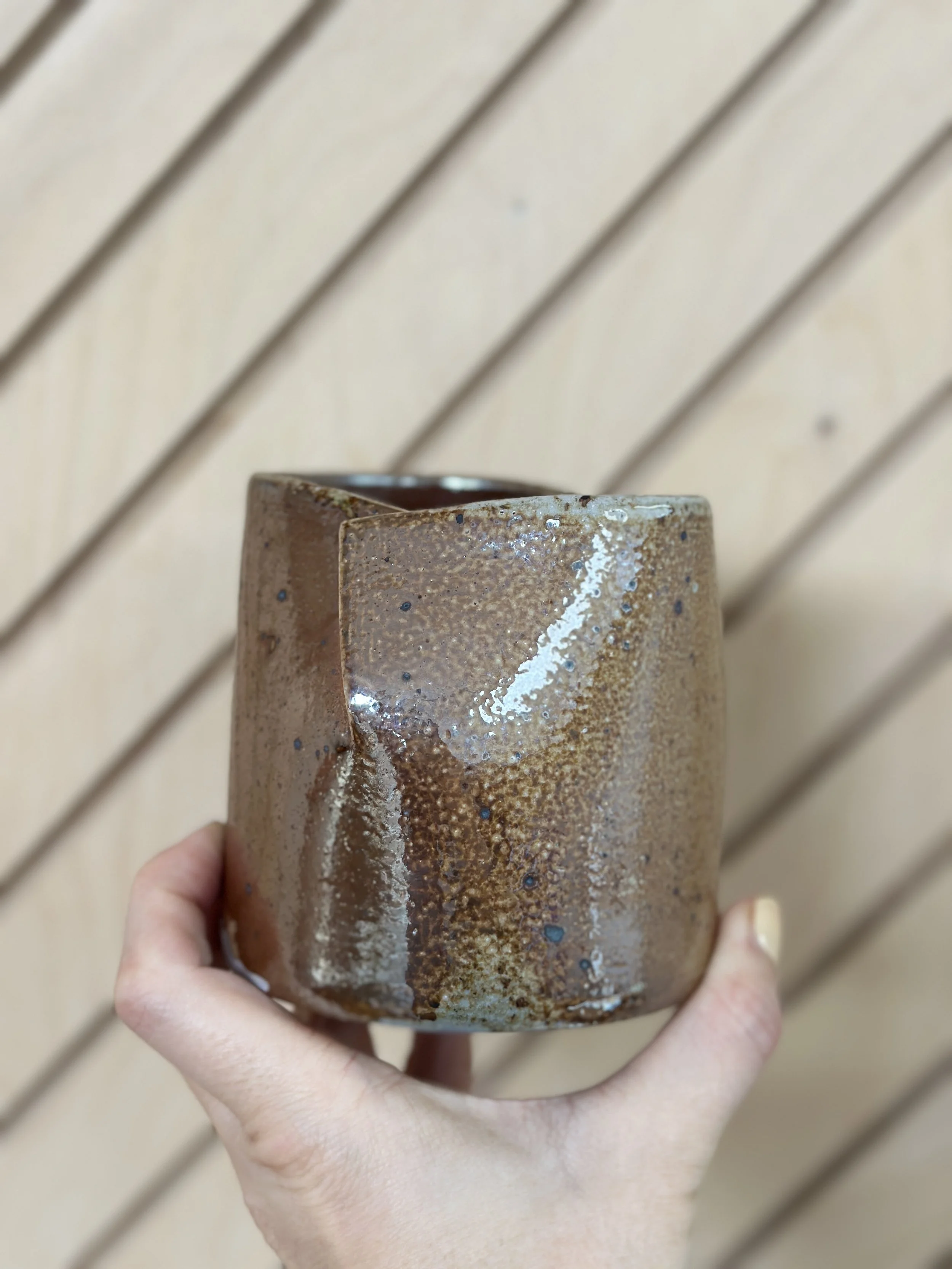 folded shiny metallic ceramic vessel held by a hand with a diagonal modern wood background