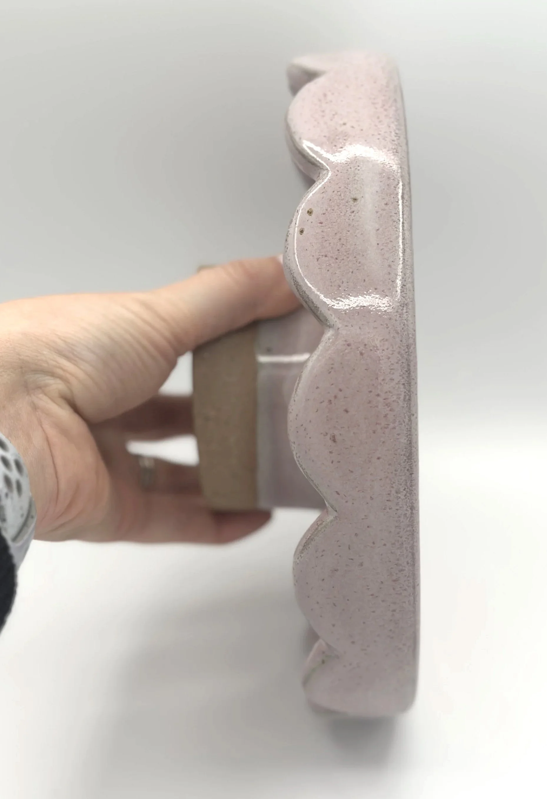 handmade stoneware ceramic cake stand with scalloped edges and pink speckled glaze held by the bottom in a hand showing off the small unglazed stem