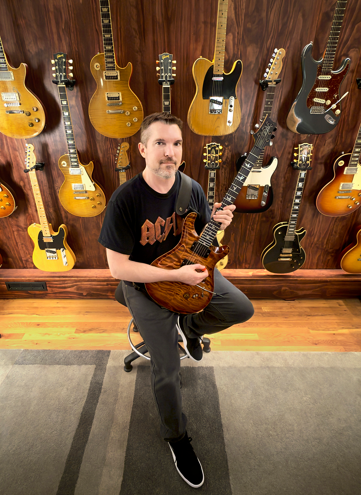 A man sitting on a stool holding an electric guitar in a music store with a wall of various guitars hanging behind him.