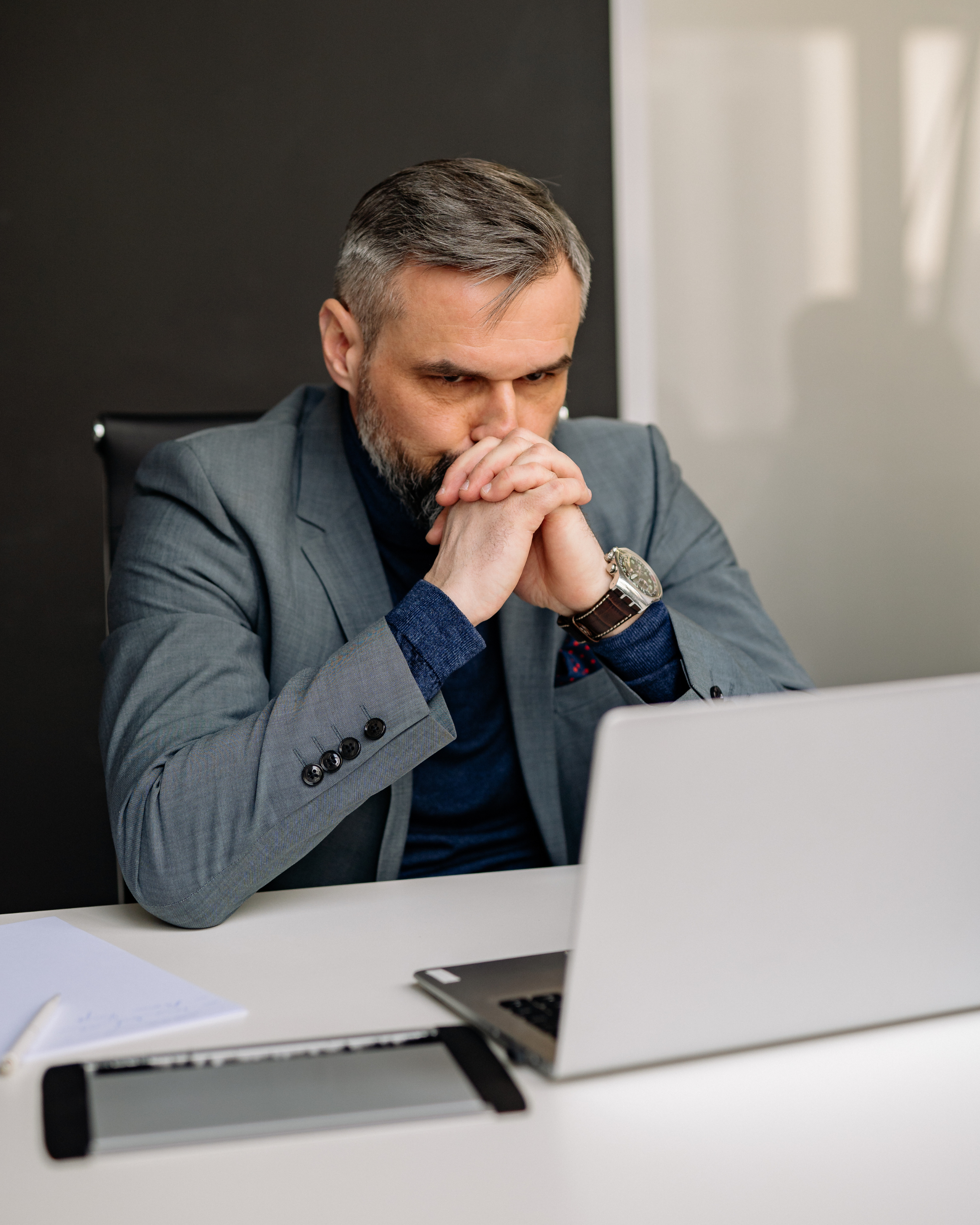 Hombre con barba y pelo gris, vistiendo saco gris y camisa azul, sentado en una oficina, con las manos juntas frente a su rostro en actitud pensativa, frente a una computadora portátil en un escritorio blanco.