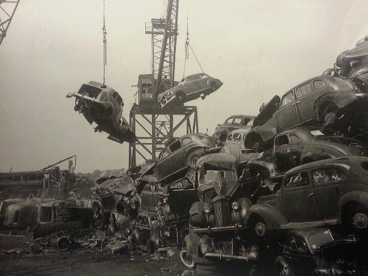 A large pile of wrecked cars stacked haphazardly beneath a crane, with some cars suspended in midair, in a scrapyard or salvage yard.