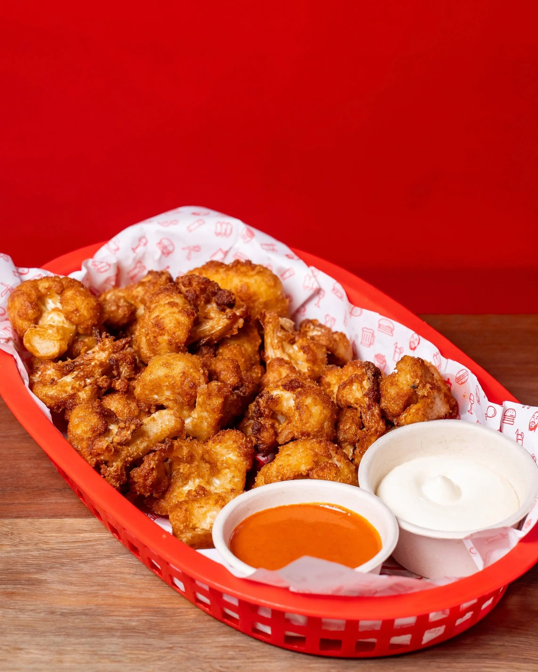 Basket of deep fried cauliflower pieces with two dipping sauces, one white and one orange (buffalo sauce and aioli), on a wooden table with a red background.