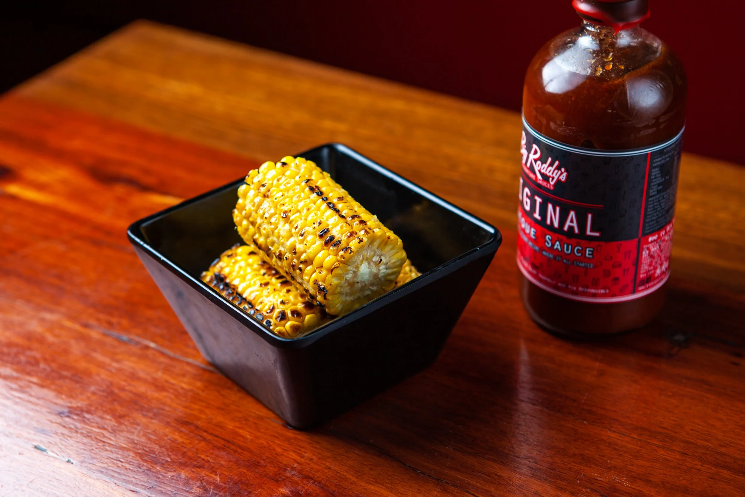 Grilled corn on the cob in a black bowl next to a bottle of barbecue sauce on a wooden table.