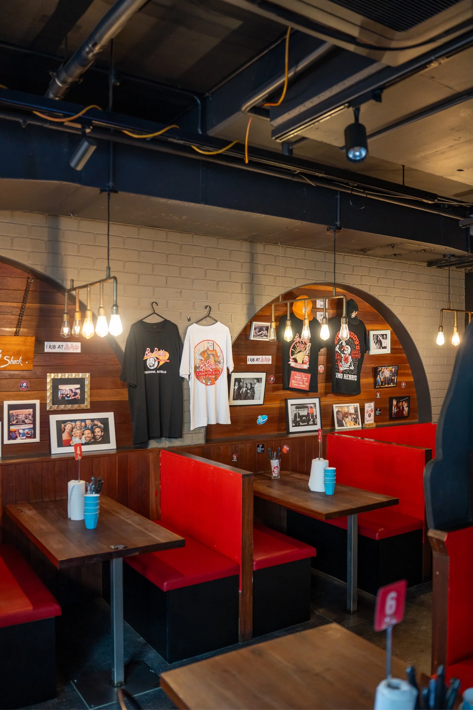 Interior of a restaurant with red booth seating (diner style), wooden (hand-crafted) tables, amusing photo decorations, and merchandise (t-shirts/ caps) hanging on the wall.