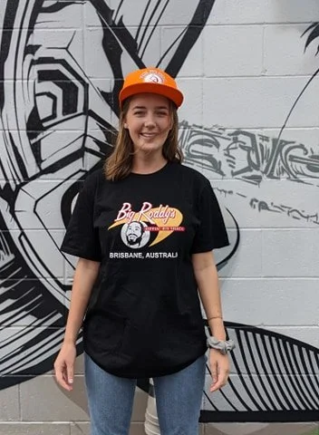 Young woman smiling, wearing Big Roddy's merchandise: An orange cap and a black t-shirt with a graphic design, standing in front of a black and white mural on a wall.
