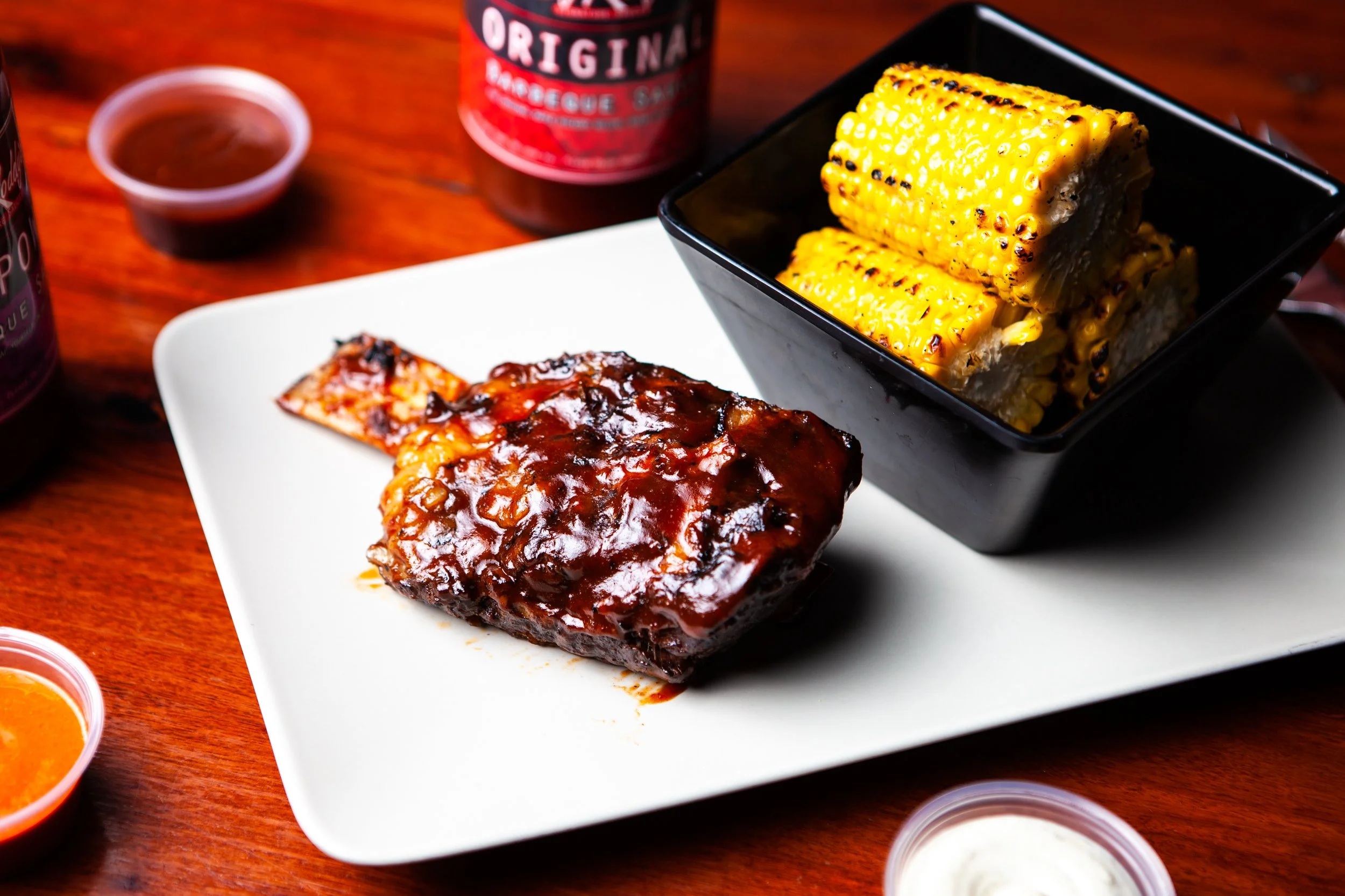 Barbecue ribs with barbecue sauce, grilled corn on the cob, and small bowls of barbecue sauce and ranch dressing on a white platter and wooden table.