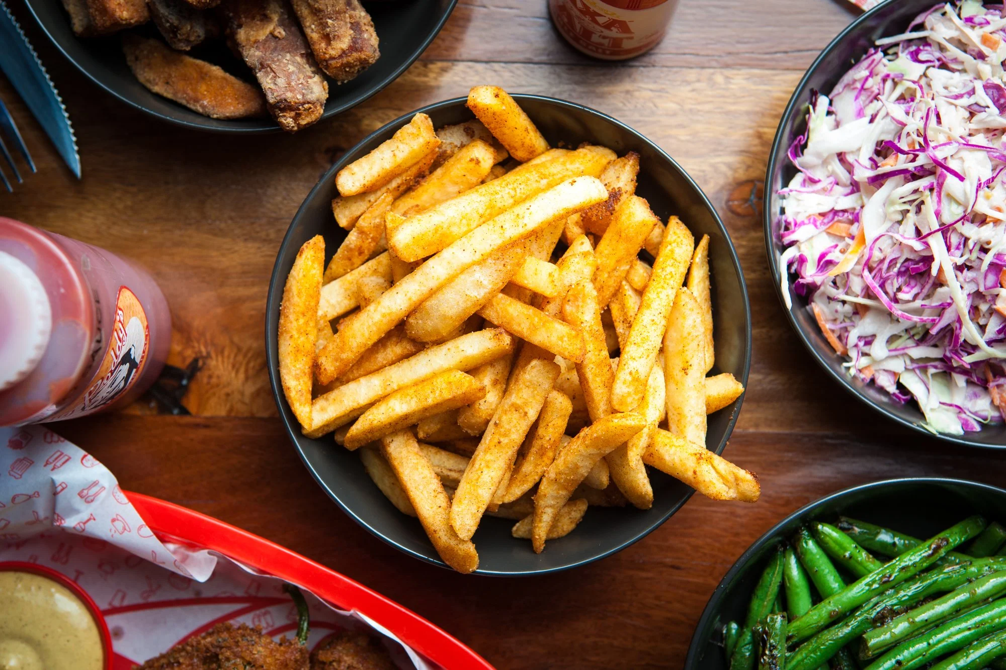 A bowl of French fries with seasoning, surrounded by coleslaw, grilled green beans, fried chicken, and a bottle of dipping sauce on a wooden table.