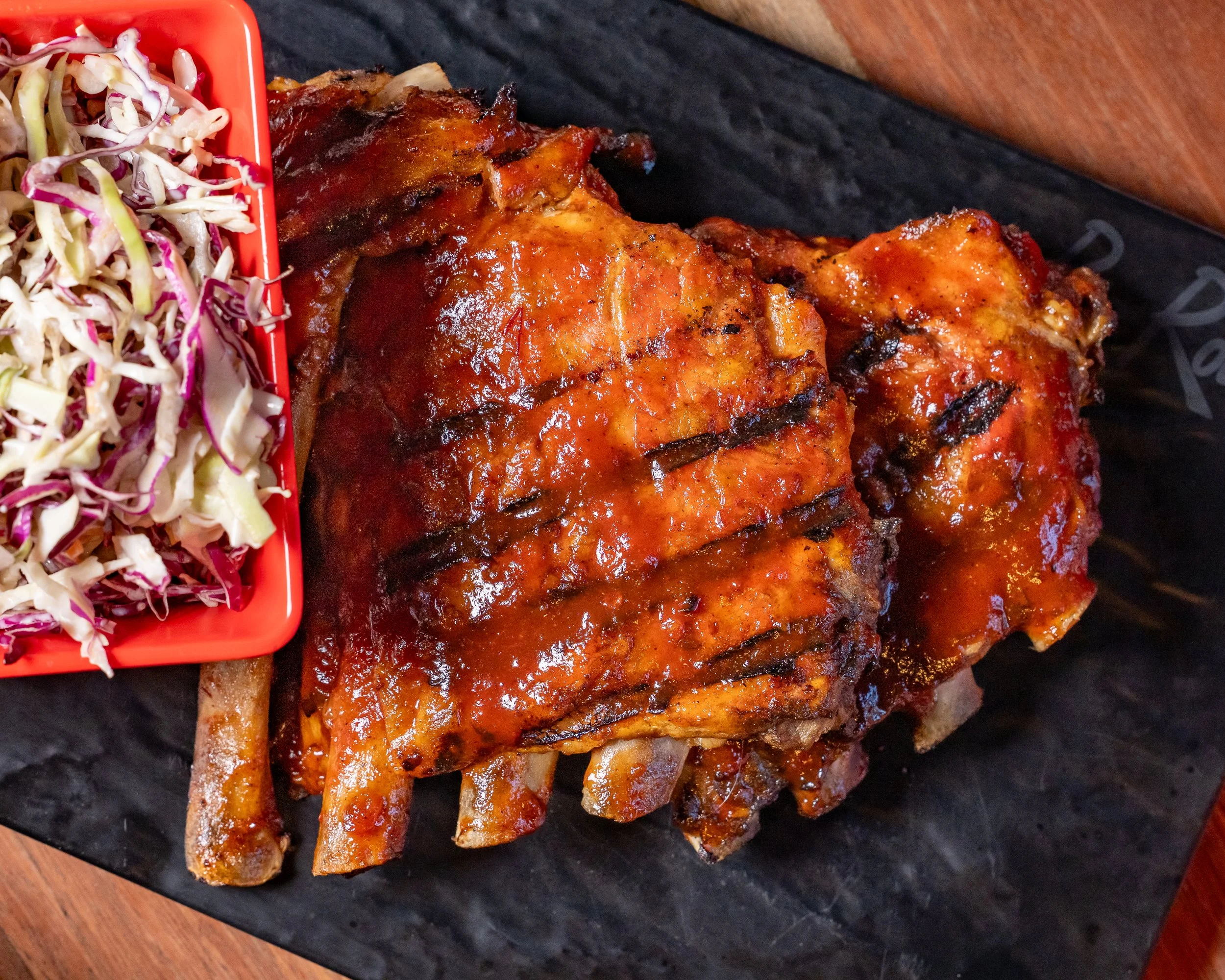 Charred barbecue pork ribs with sauce next to a side of coleslaw in a red dish on a black serving board.