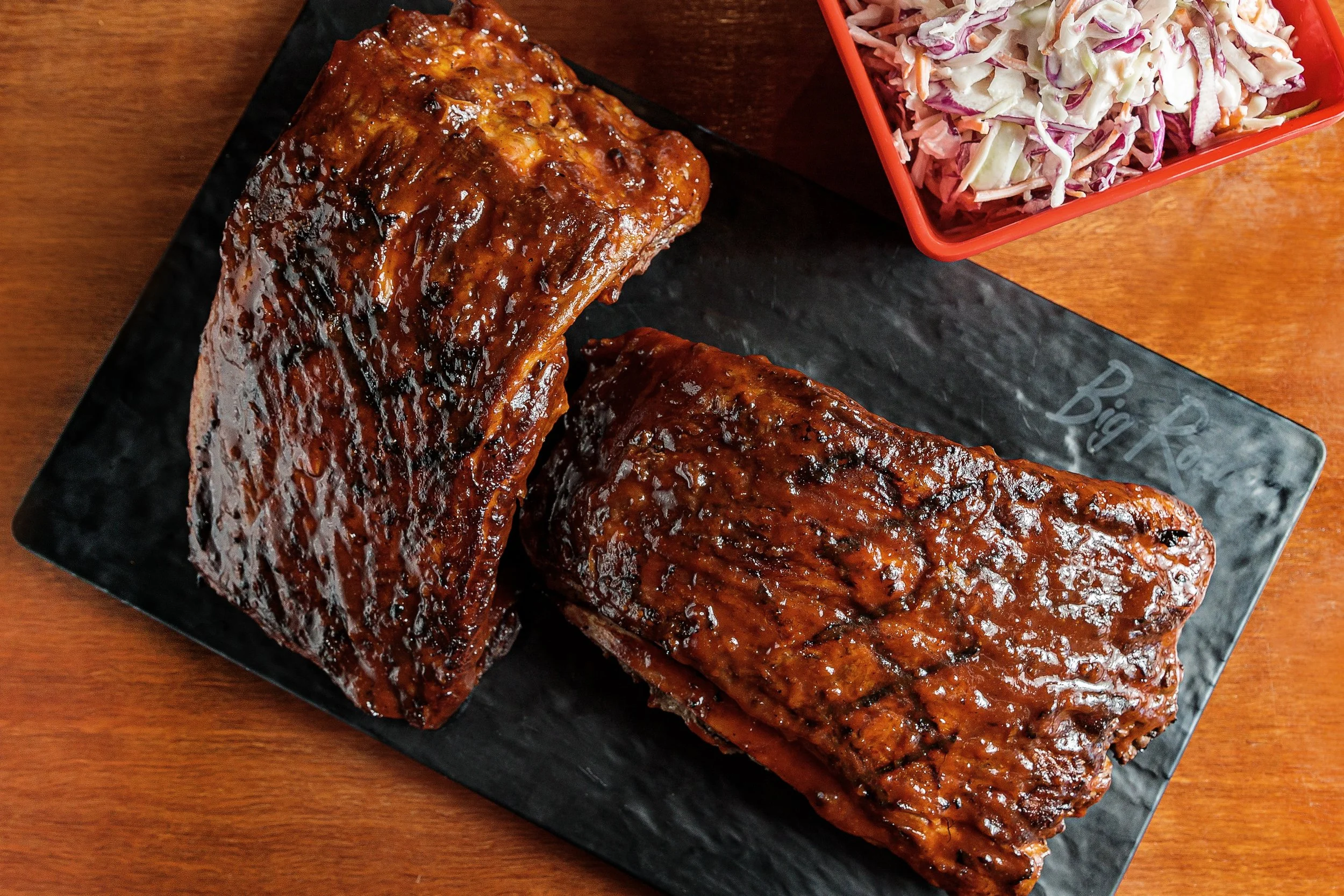 Two glazed racks of barbecue ribs on a black plate with a side of coleslaw in a red container, on a wooden table.