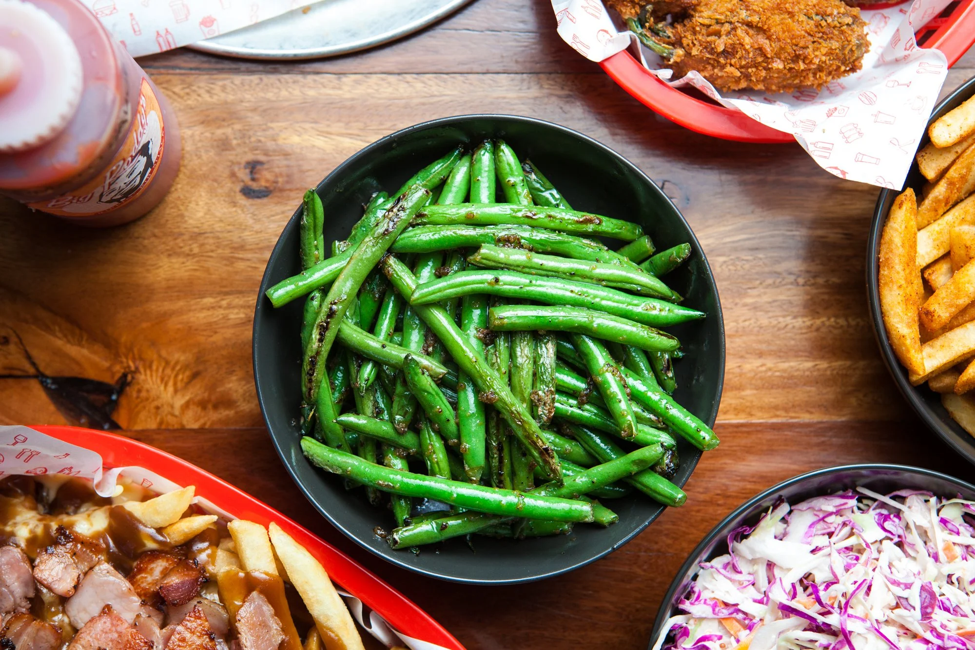 A black bowl of cooked garlic-pan fried green beans on a wooden table, surrounded by other food dishes including fried chicken, French fries, coleslaw, and a bottle of barbecue sauce.
