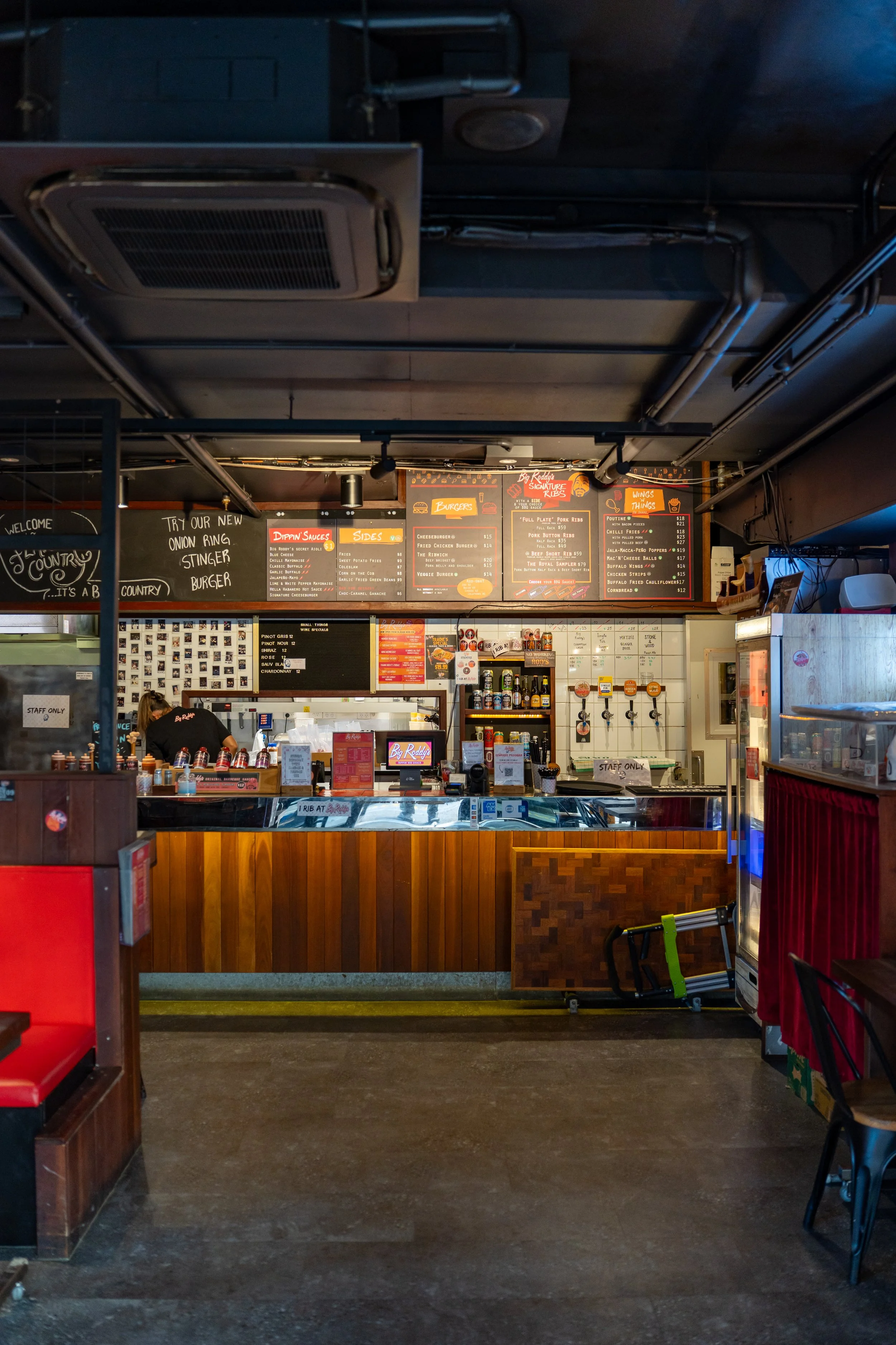 Interior of a casual burger restaurant with a front counter, bar with taps of draft beer, menus on blackboards, and seating area with red booths, American diner style.