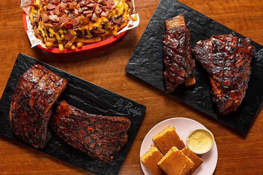 A spread of barbecue ribs (pork ribs and beef ribs), authentic poutine with crispy bacon, and cornbread with maple butter on a wooden table.