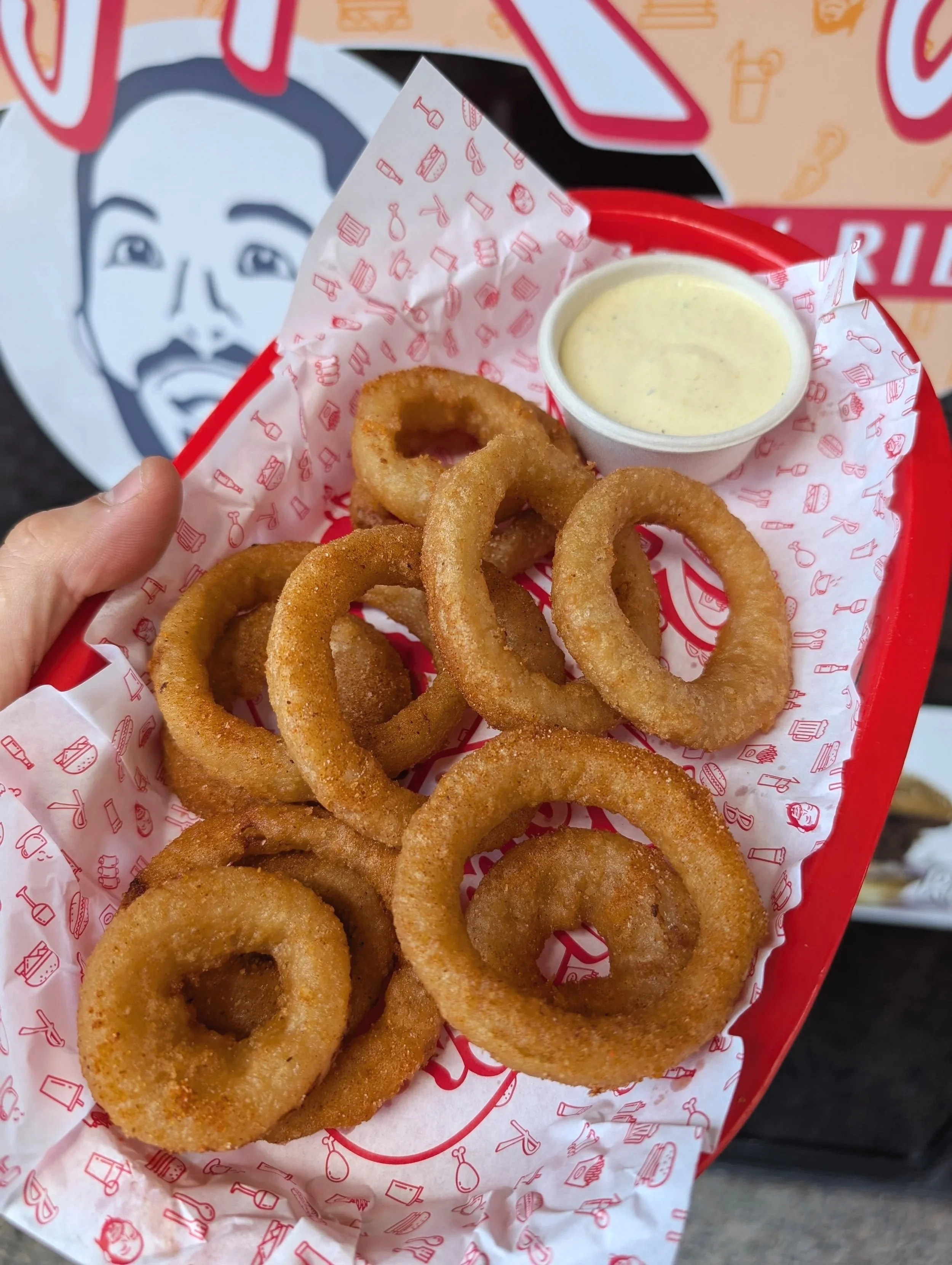 A red basket of crispy fried onion rings with a side of creamy dipping sauce on red and white patterned paper, with a smiling man and a wall with signs in the background.