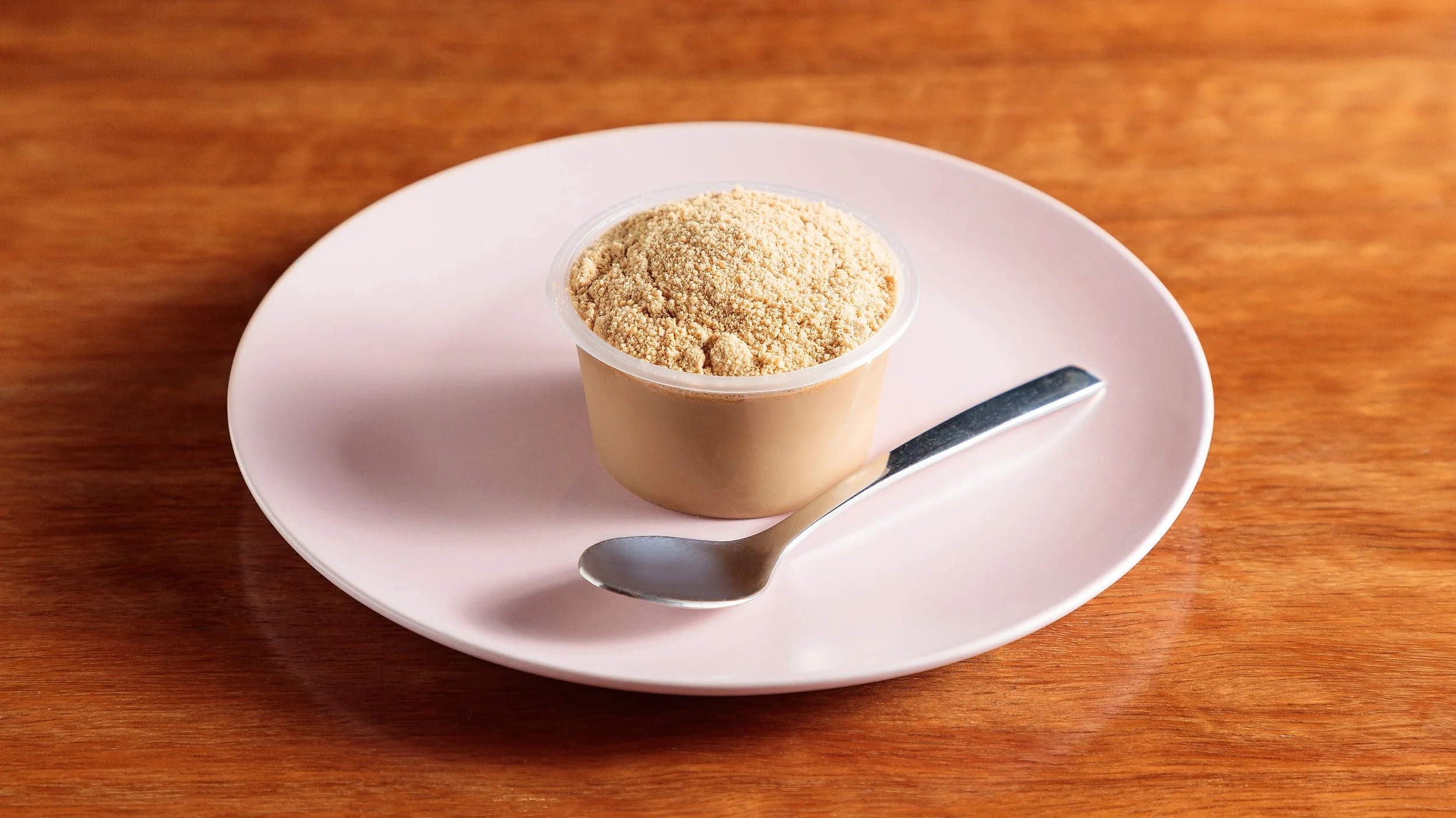 A cup of chocolate ganache moose with cookie crumb on a pink plate with a small spoon, placed on a wooden surface.
