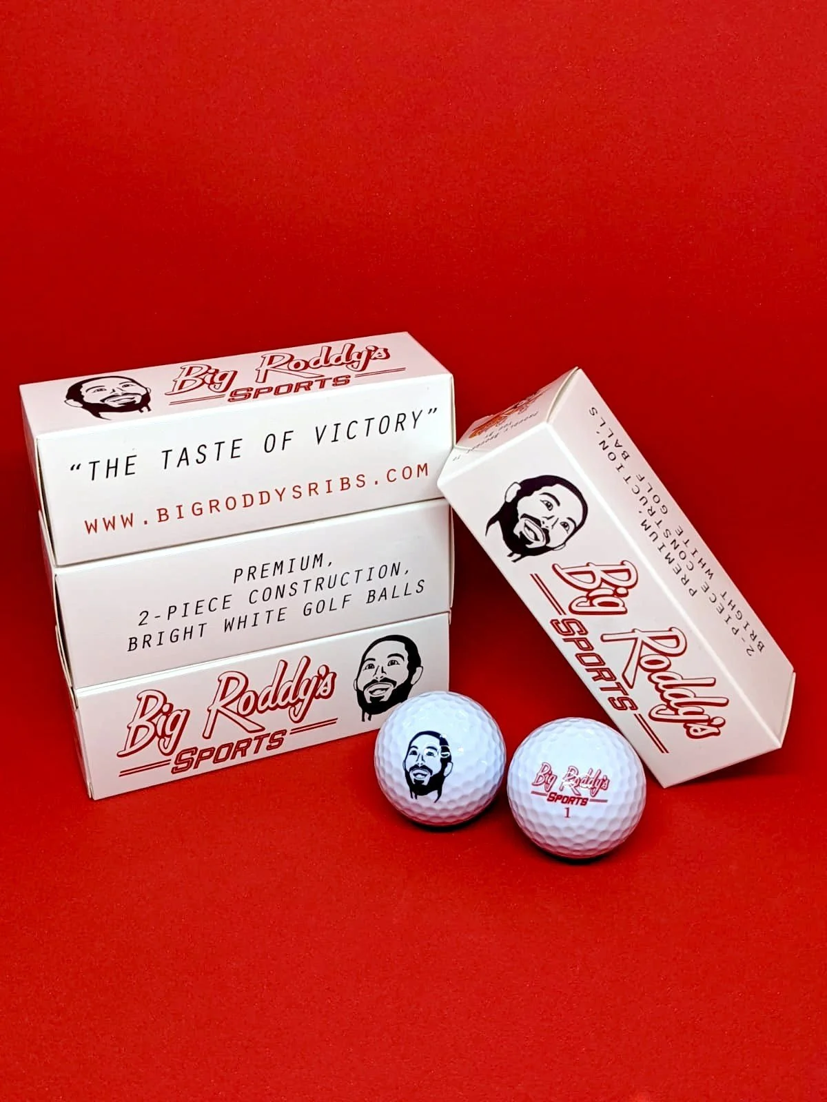Two white golf balls with the Big Roddys Sports logo and a cartoon man's face, along with a box and a pack of golf balls from Big Roddys Sports on a red background.