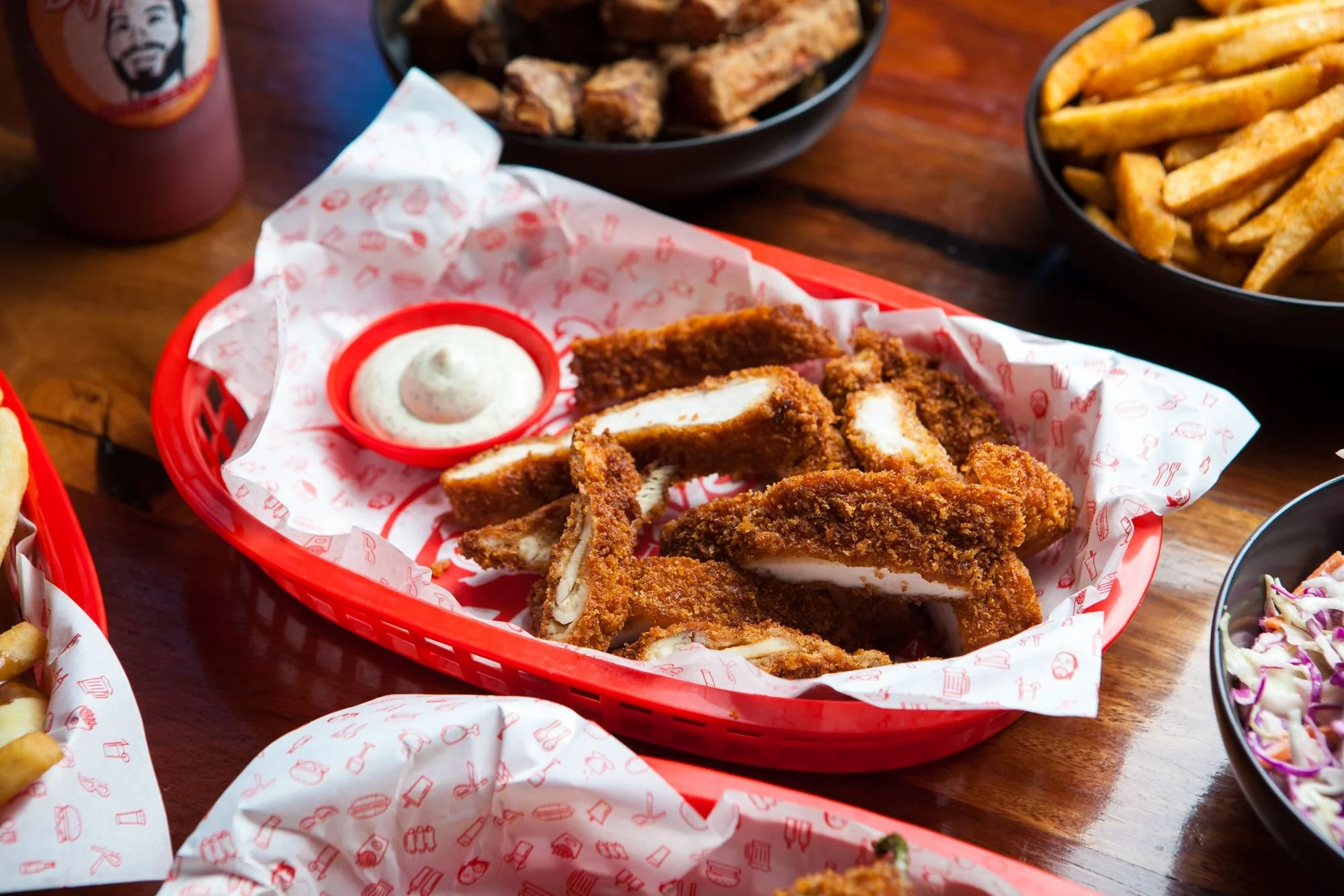 Fried chicken breast tenders/ strips with a side of ranch dipping sauce on a red basket liner, surrounded by bowls of fries and coleslaw on a wooden table.