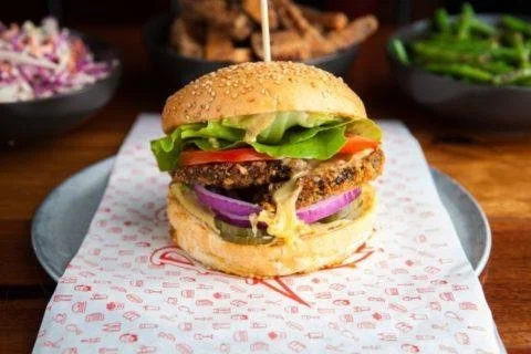 Juicy burger with lettuce, tomato, onions, cheese, and a beef patty, served on a sesame seed bun with side dishes in background.