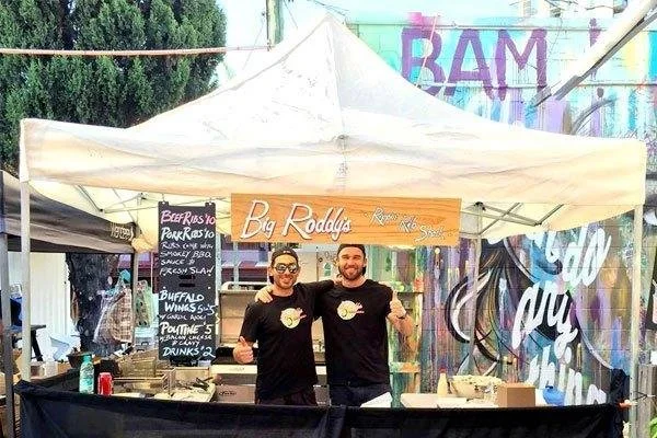 Two men standing behind a food stall with a white canopy at a market, smiling and giving thumbs up. The stall has a sign that reads 'Big Roddy's' and a menu board listing various foods including ribs (beef, pork), buffalo wings and poutine (fries).