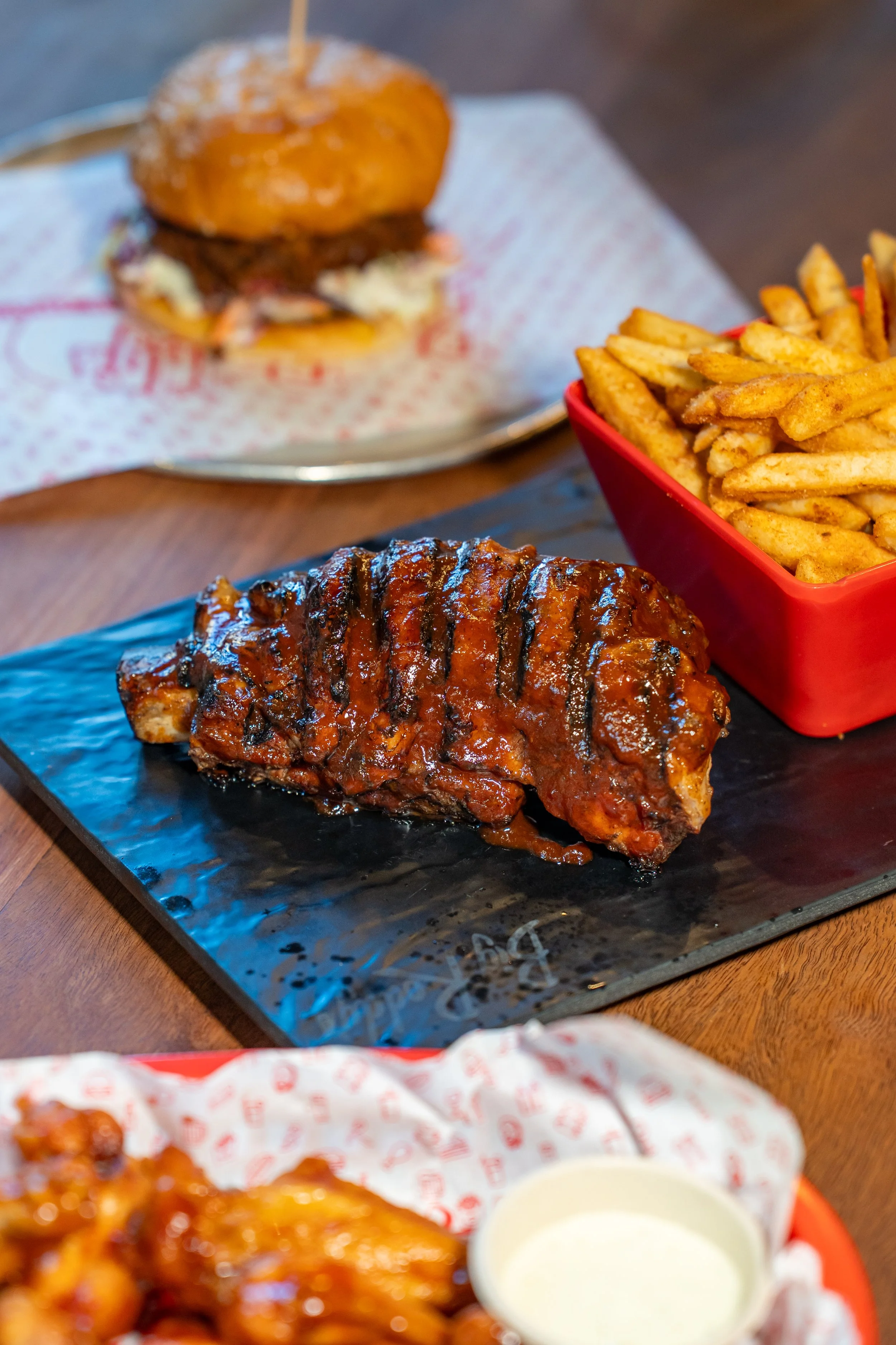 Beef Ribs served on a black slate platter with grilled markings from the grill. Side of French fries in a red container, and a blurred background with a pulled meat burger on a plate, with a small cup of white dipping sauce in the foreground.