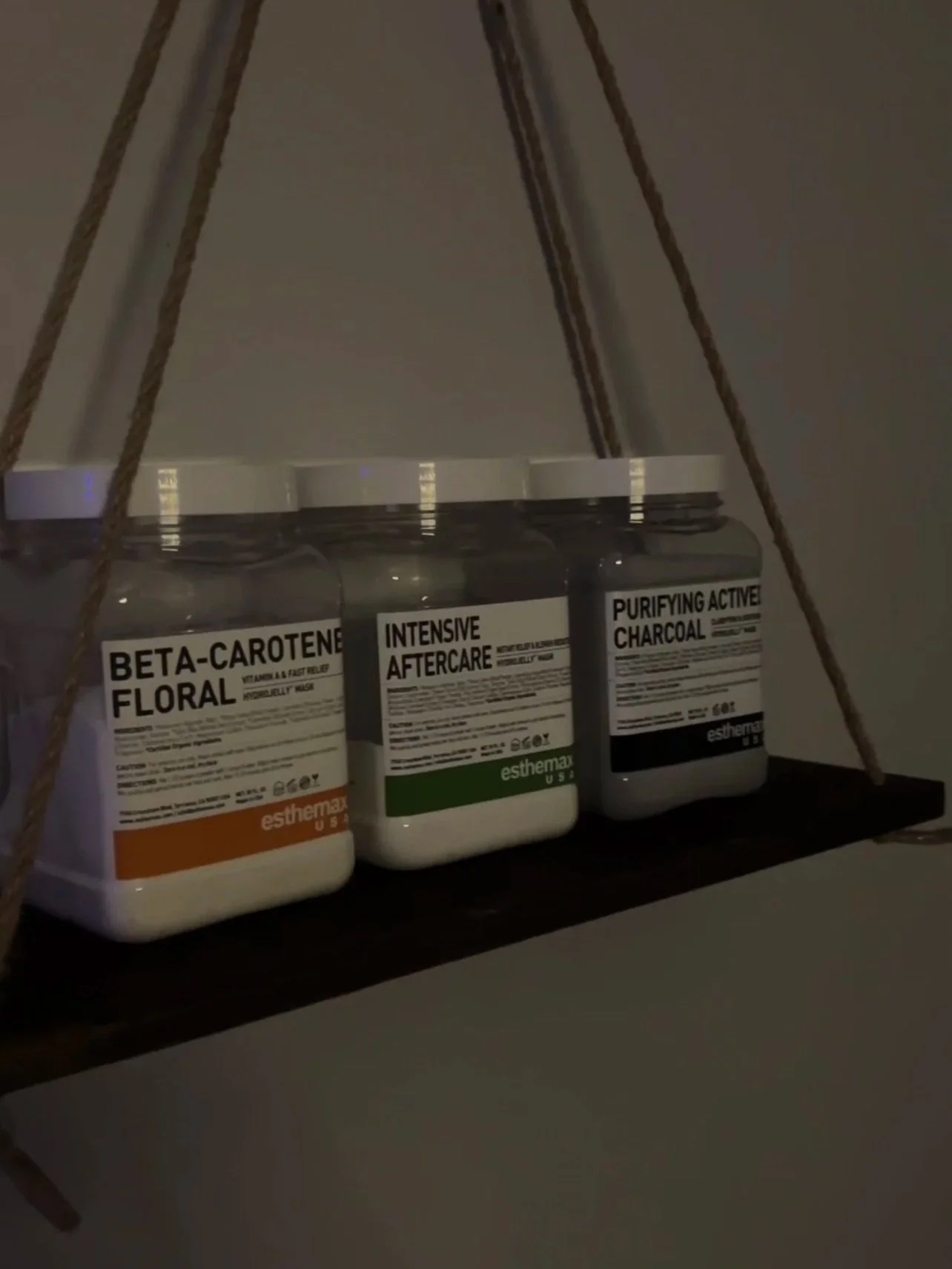Three jars of supplements labeled Beta-Carotene Floral, Intensive Aftercare, and Purifying Active Charcoal on a dark wooden shelf with ropes holding it up.