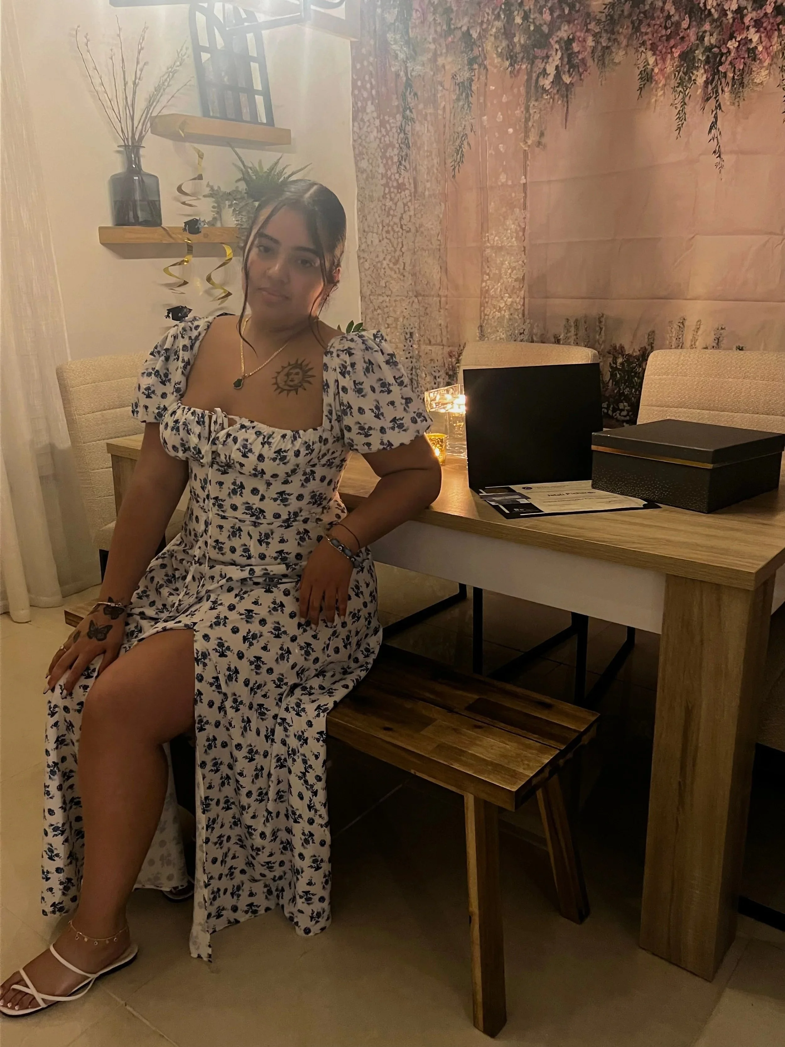A young woman sitting on a wooden bench in a dining room, wearing a white floral dress with puffed sleeves, sandals, and jewelry, with a tattoo of a sun on her chest. Behind her, there's a wooden table with black gift boxes, a lit candle, and a dark laptop. The background features shelves with decorative items and a pink floral wall.