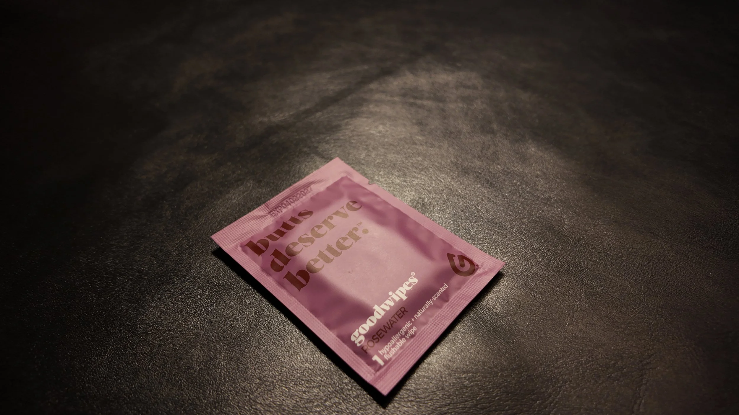 A pink packet of wet wipes labeled 'Just Deserve Better' with the scent rosewater, lying on a dark textured surface.