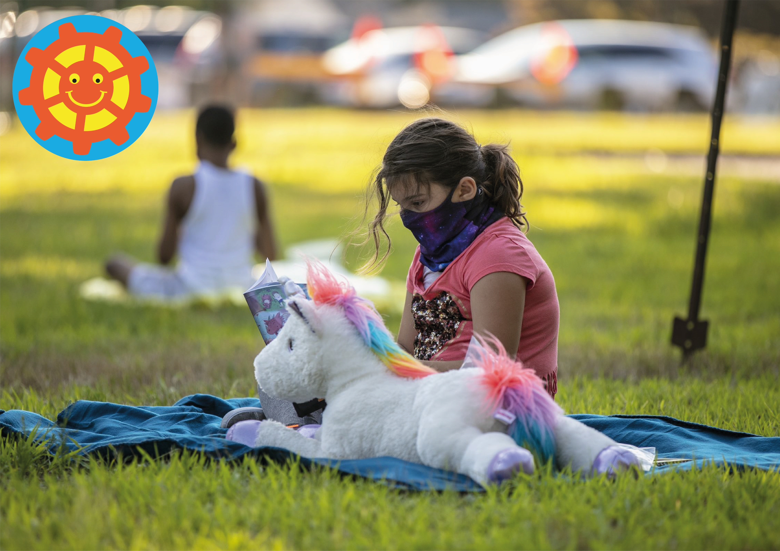 A girl sits on a blanket outdoors, reading a book, with a rainbow-colored stuffed unicorn toy beside her.