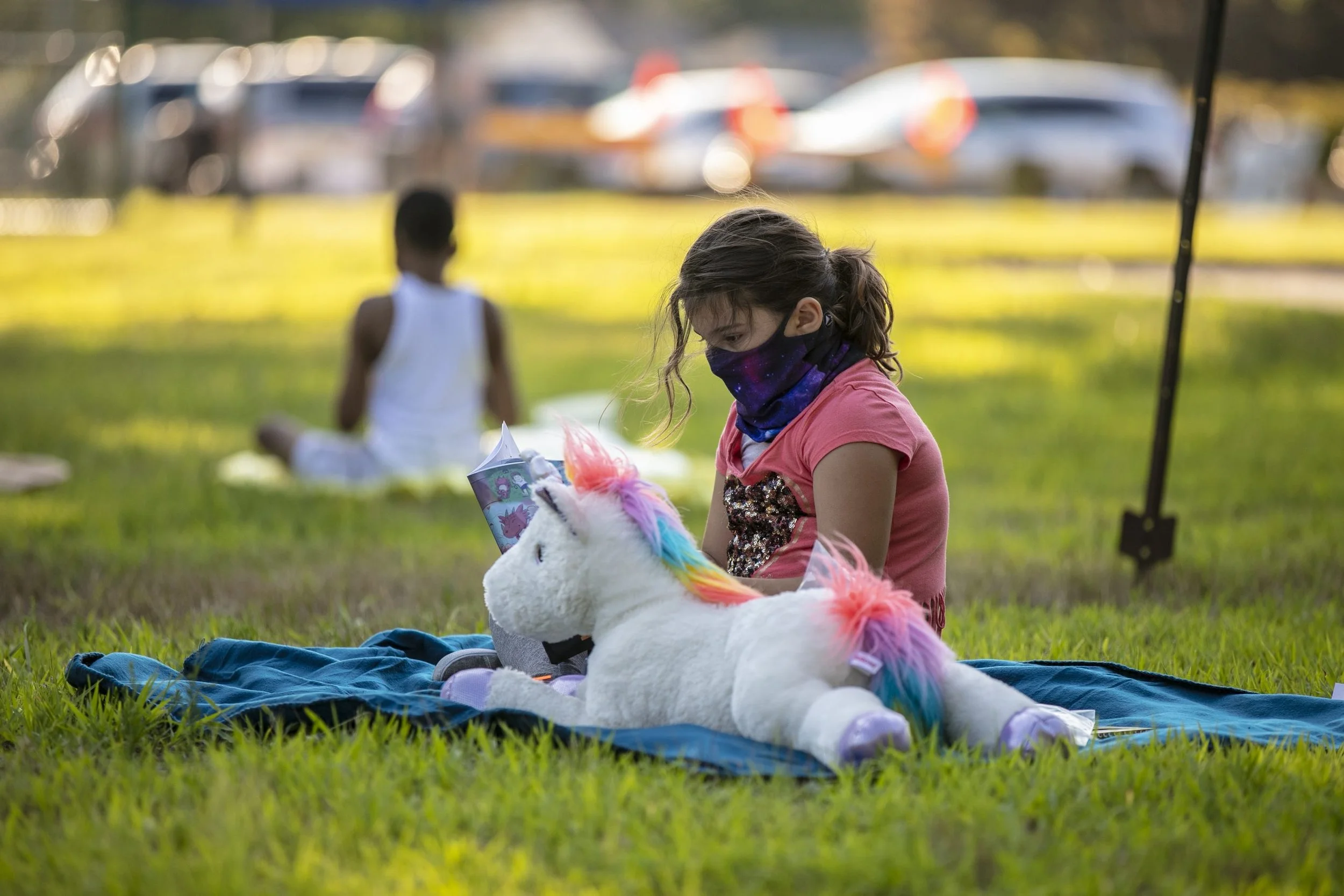 A young girl sitting on a blue blanket in a grassy park, reading a book beside a large white plush unicorn toy with rainbow-colored mane and tail.
