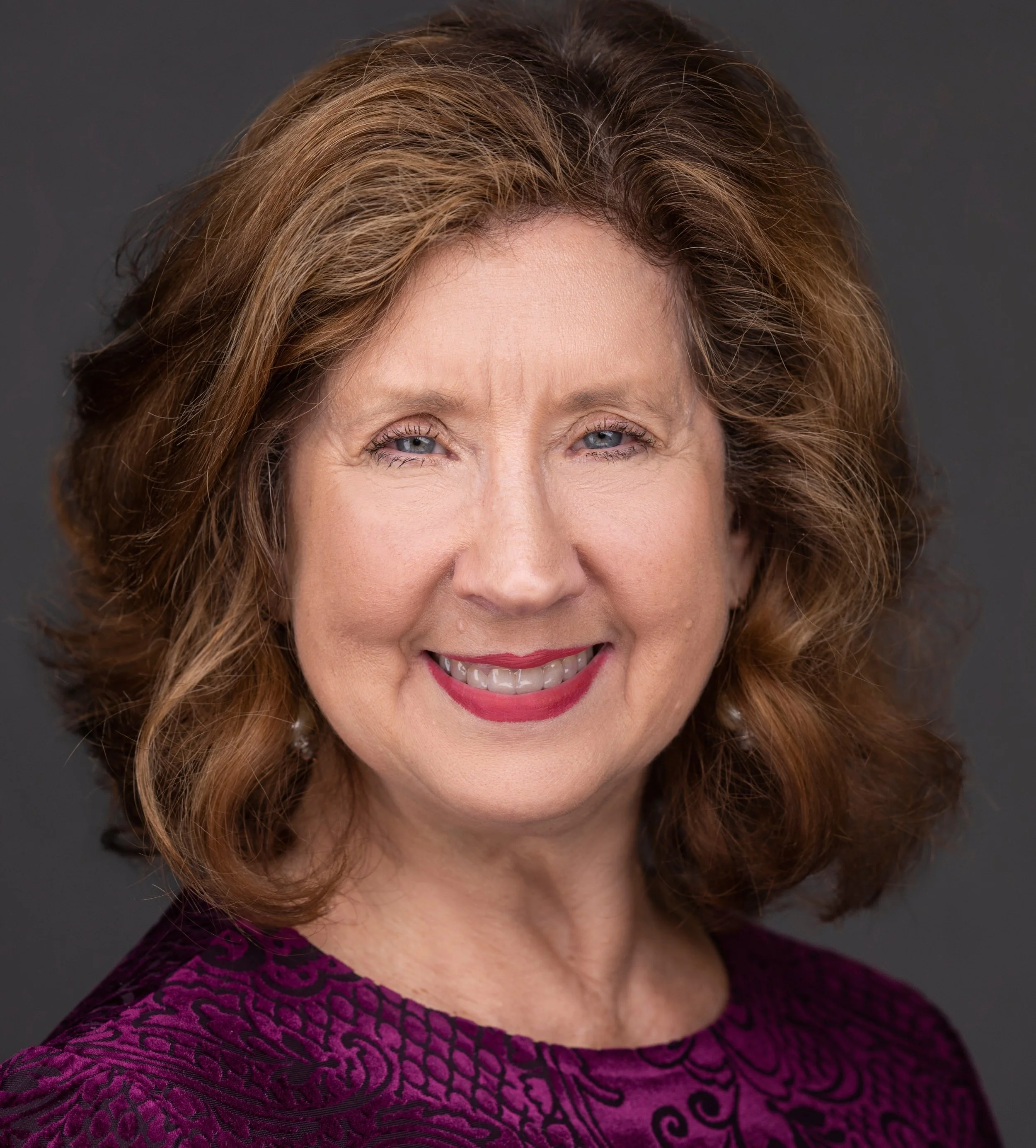 Close-up of a woman with light brown, curly hair, blue eyes, and wearing a purple velvet dress with black floral pattern, against a gray background.
