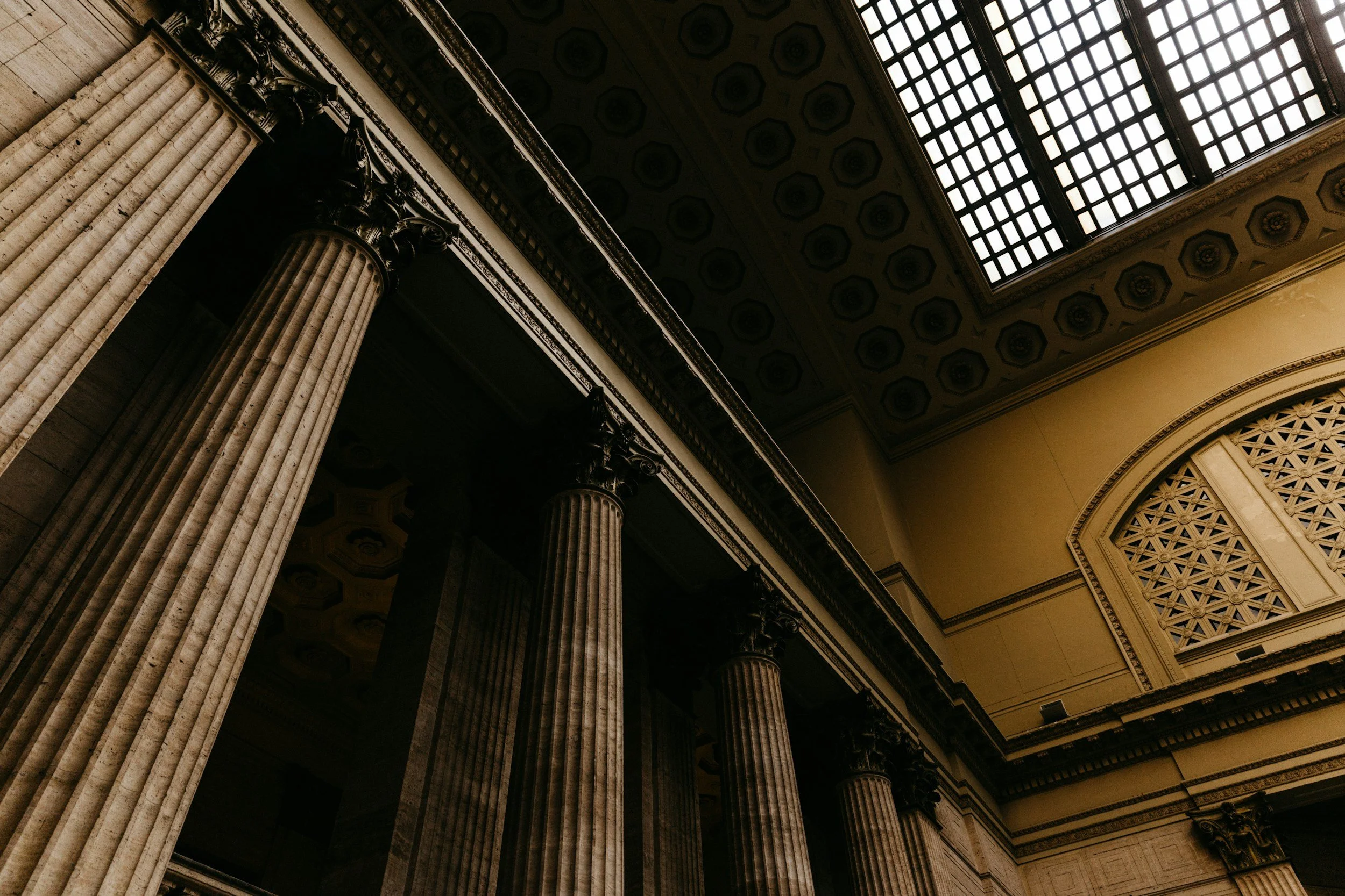 Interior ceiling view of a grand building with large columns, decorative ceiling, and a large skylight allowing natural light to illuminate the space.