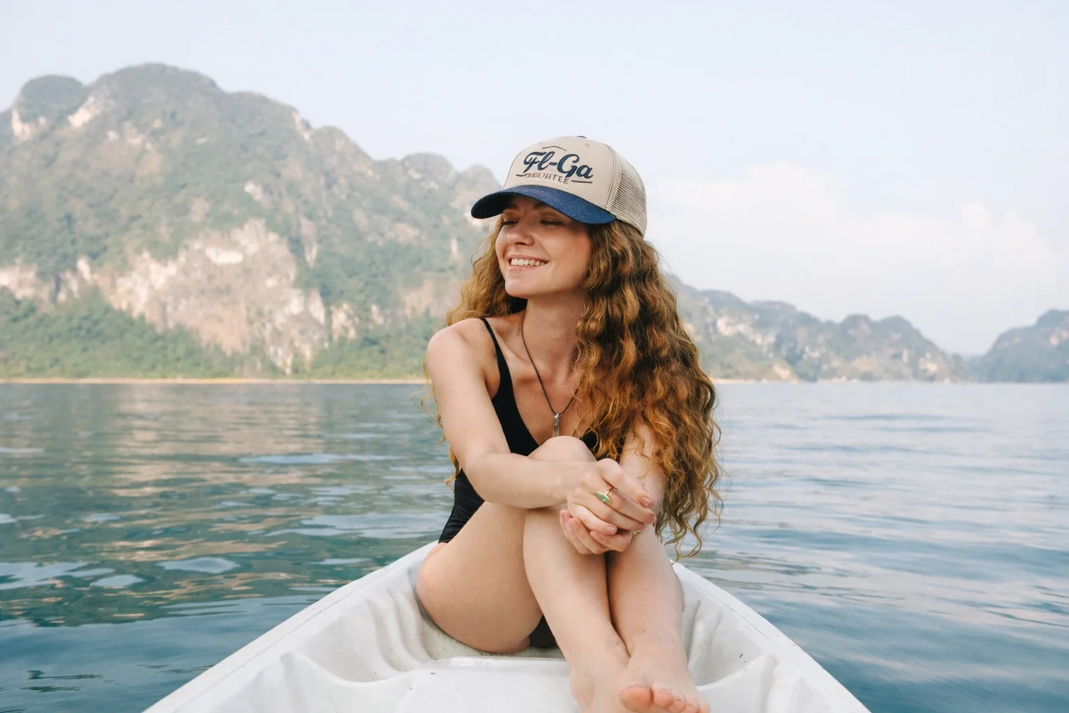 A young woman with curly red hair, wearing a trucker cap, black swimsuit, and jewelry, sitting in a white boat on calm water with lush, green hills and mountains in the background.