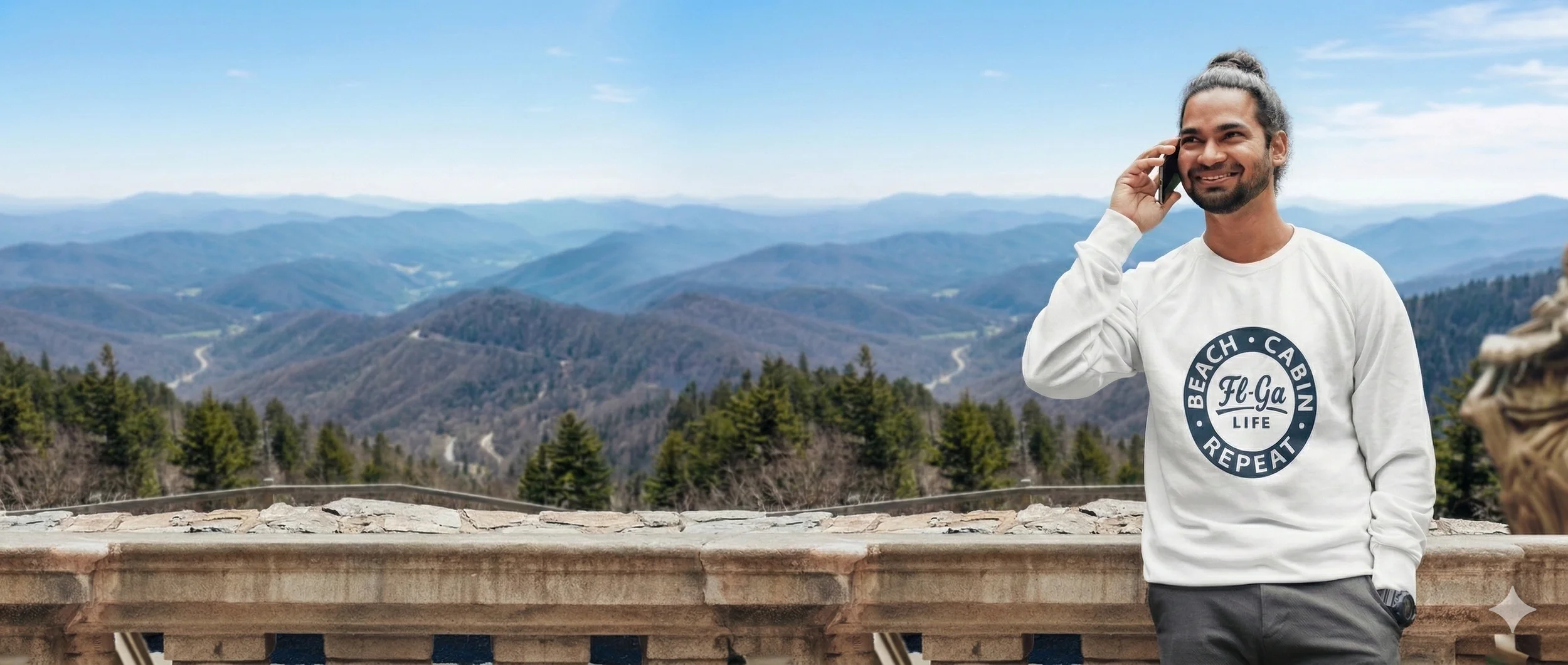 Man standing on a balcony, talking on a cellphone with mountains and forests in the background, wearing a white sweatshirt with a circular logo that reads 'Beach Cabina Repeat Fl-Ga Life'.
