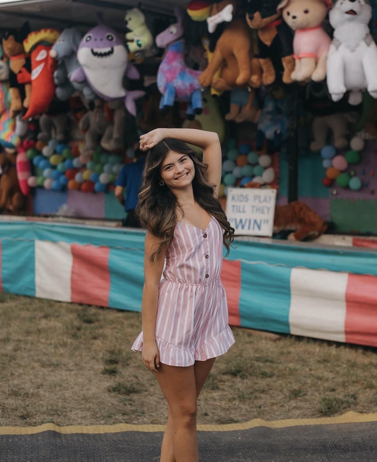 Young woman with long wavy brown hair smiling at a carnival game booth featuring plush toys and colorful decorations.
