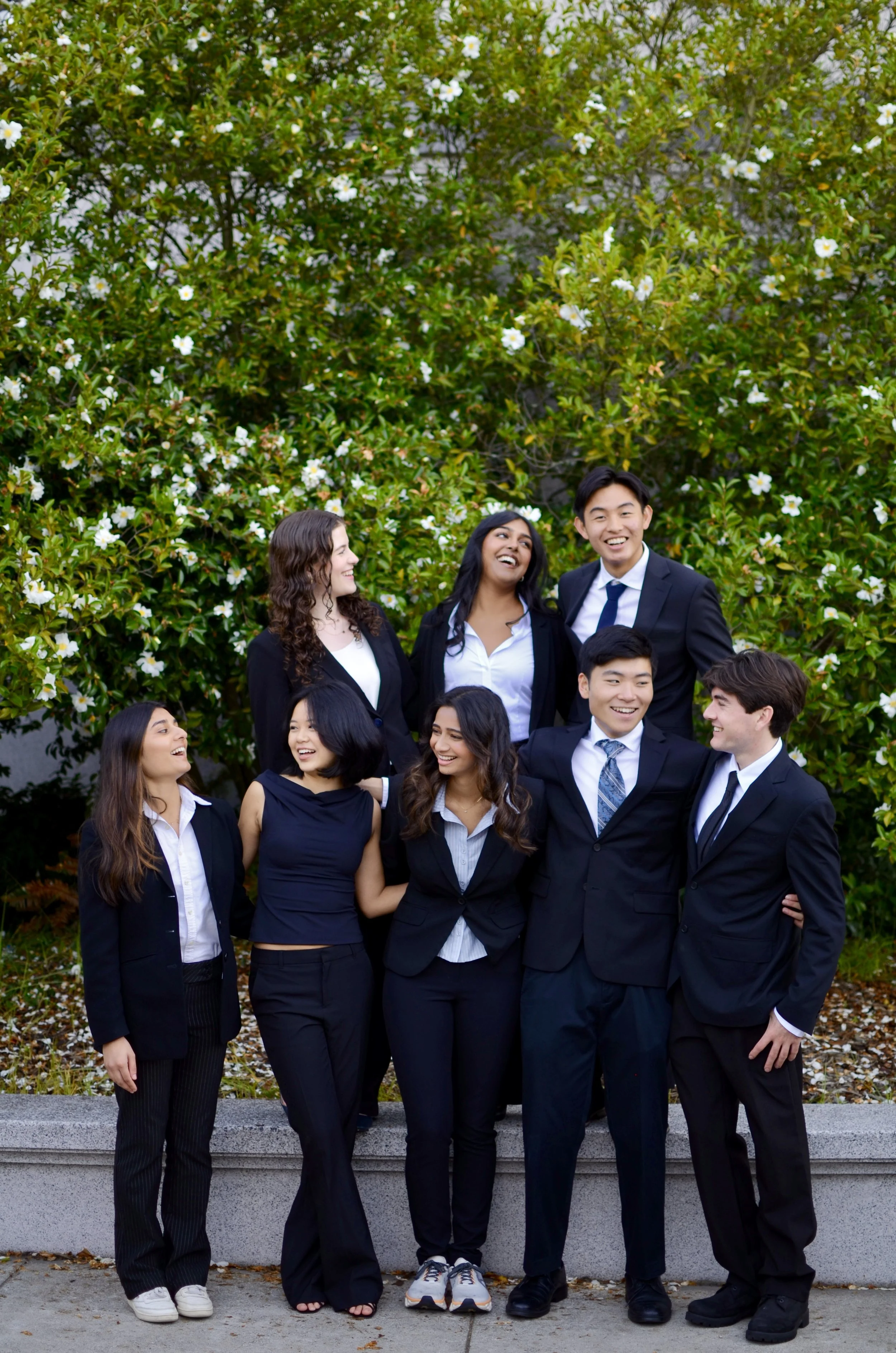 Group of seven diverse young professionals in business attire standing outdoors in front of a flowering bush, smiling and engaging with each other.