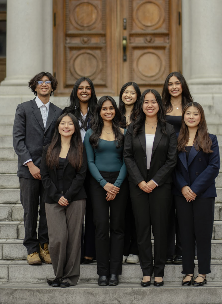 Group of nine young professionals standing on the steps of a building with large wooden doors, smiling at the camera.