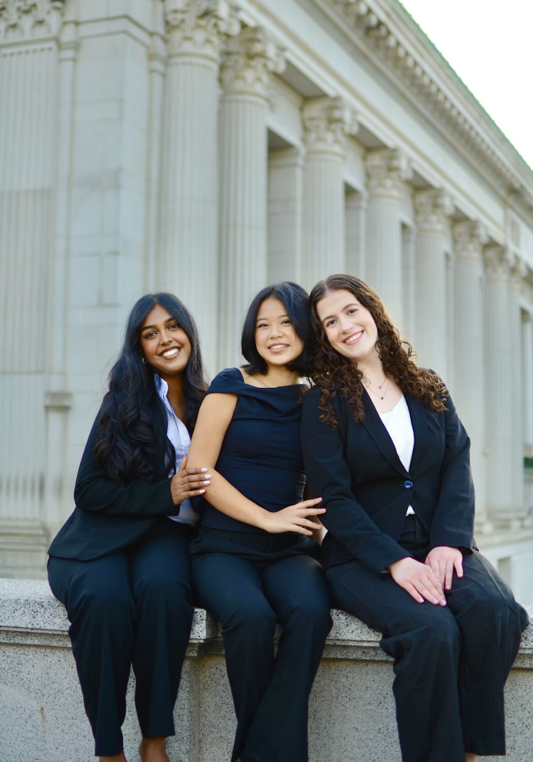 Three women sitting on a ledge outside a building with classical columns, dressed in business formal attire, smiling at the camera.