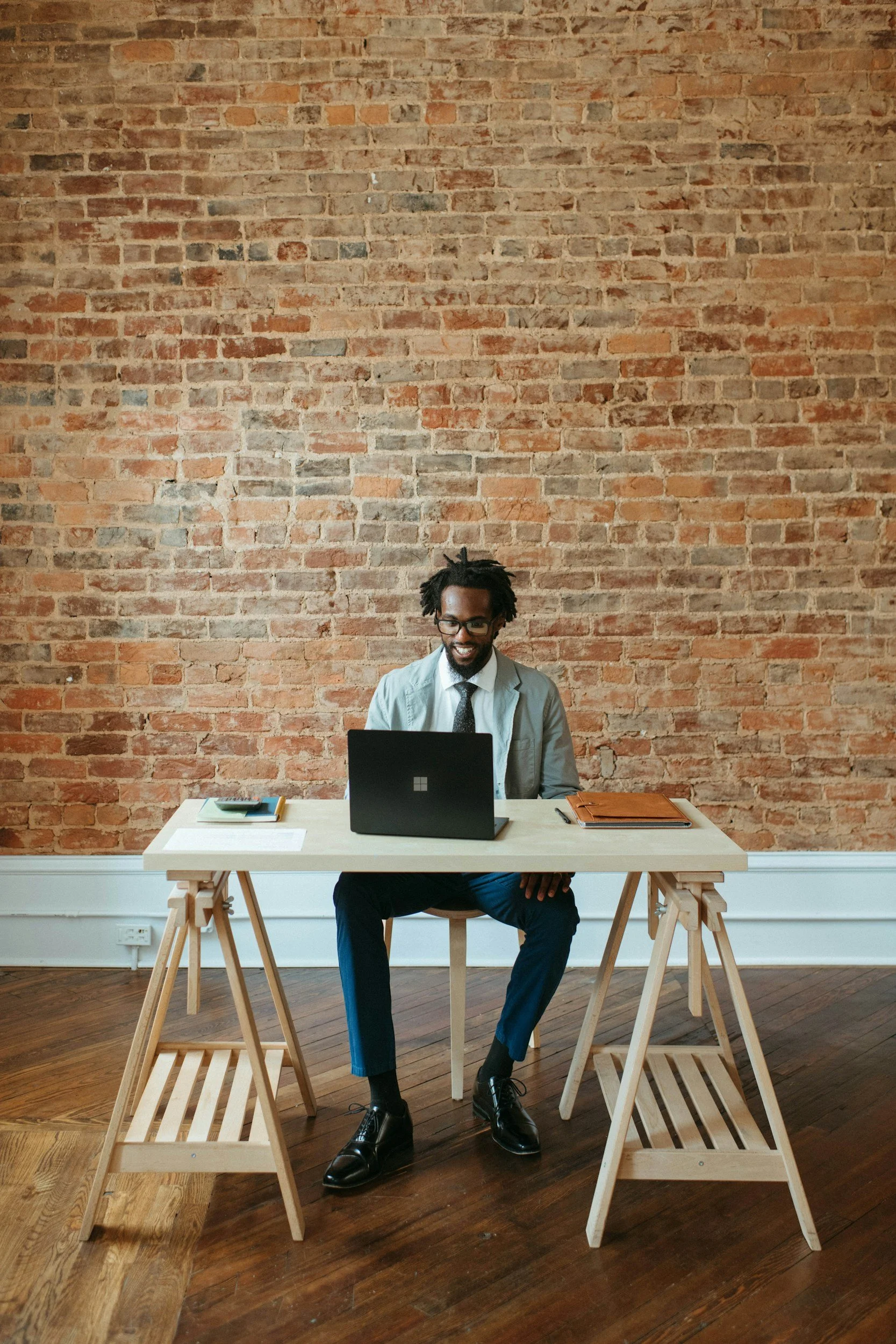 Man in professional attire sitting at a desk with a laptop, notebooks, and papers, smiling, in an office with a brick wall background.