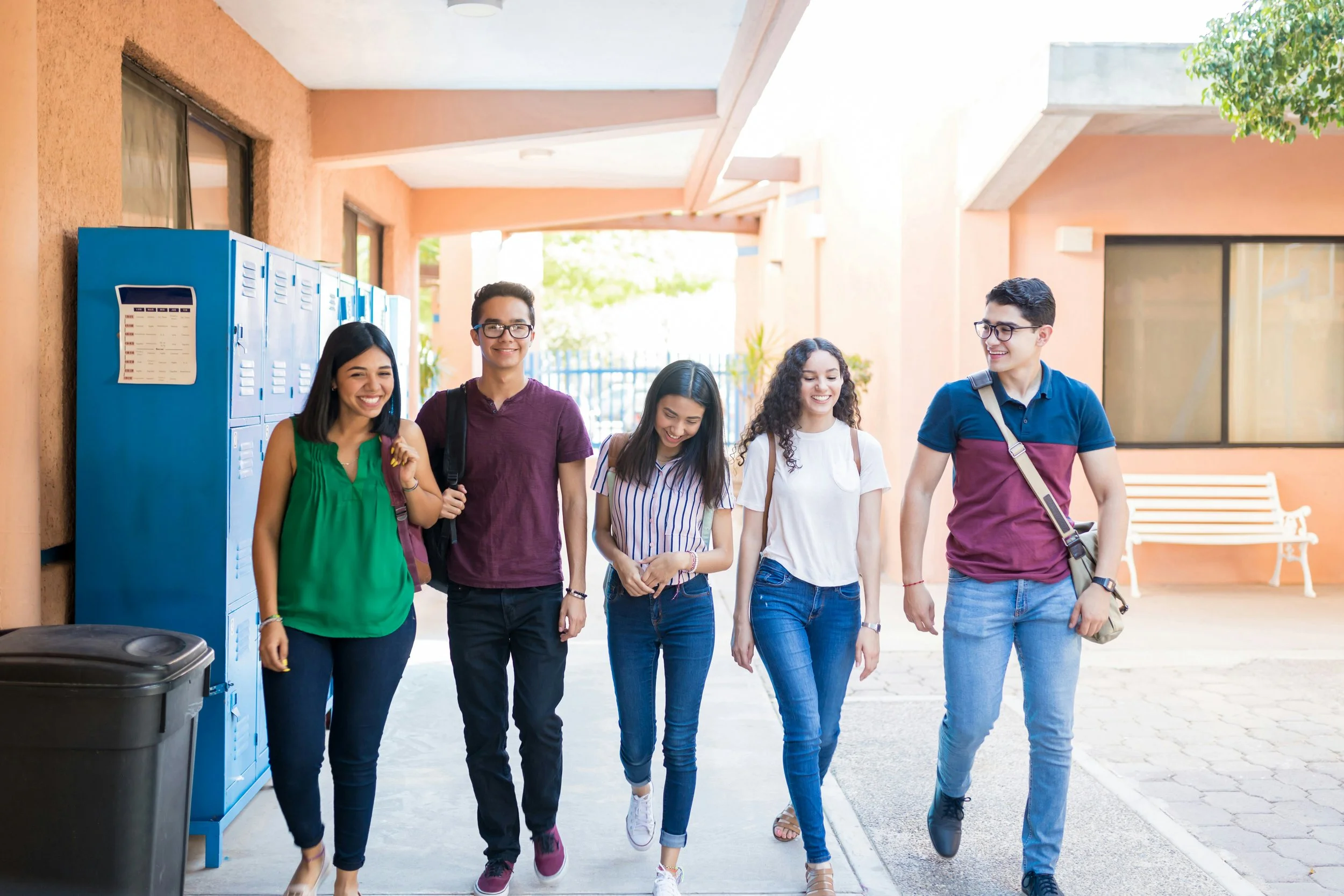 Group of five teenagers walking together in a school corridor with lockers, smiling and talking.
