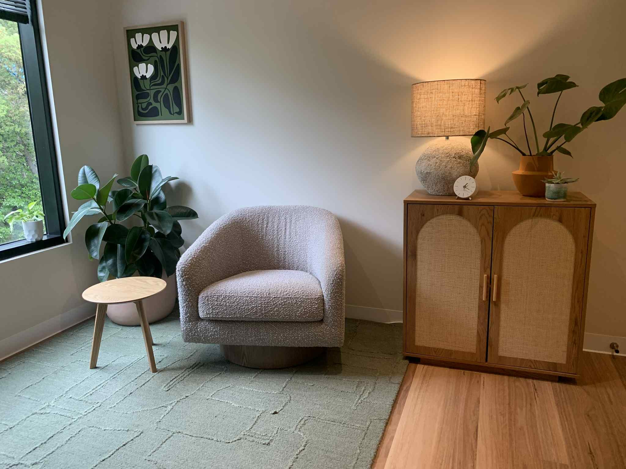 A cozy corner with a beige armchair, small wooden side table, green plant near the window, and a wooden cabinet with a lamp, pottery, and small decorations, illuminated by warm light.