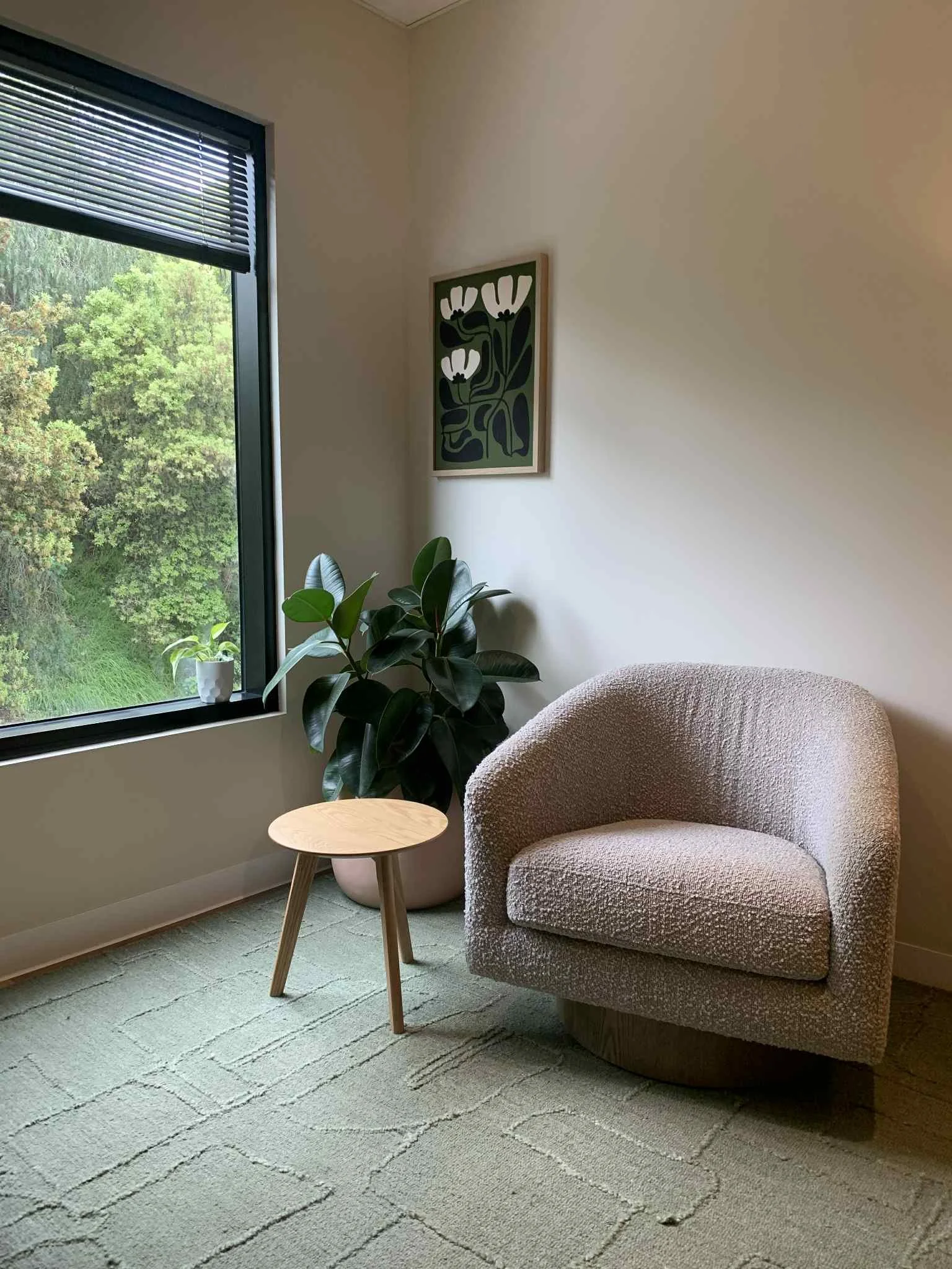 Corner of a room with beige carpet, a round armchair upholstered in textured fabric, tall leafy houseplant in a pink pot, small wooden side table, and a window with blinds overlooking greenery outside, with a framed floral art print on the wall.
