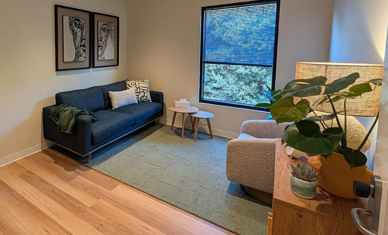 A cozy therapy room with a dark blue sofa, framed wall art, two small wooden side tables, a beige armchair, a large window with blinds, a green area rug, a wooden cabinet with potted plants, and a beautiful leafy window view.