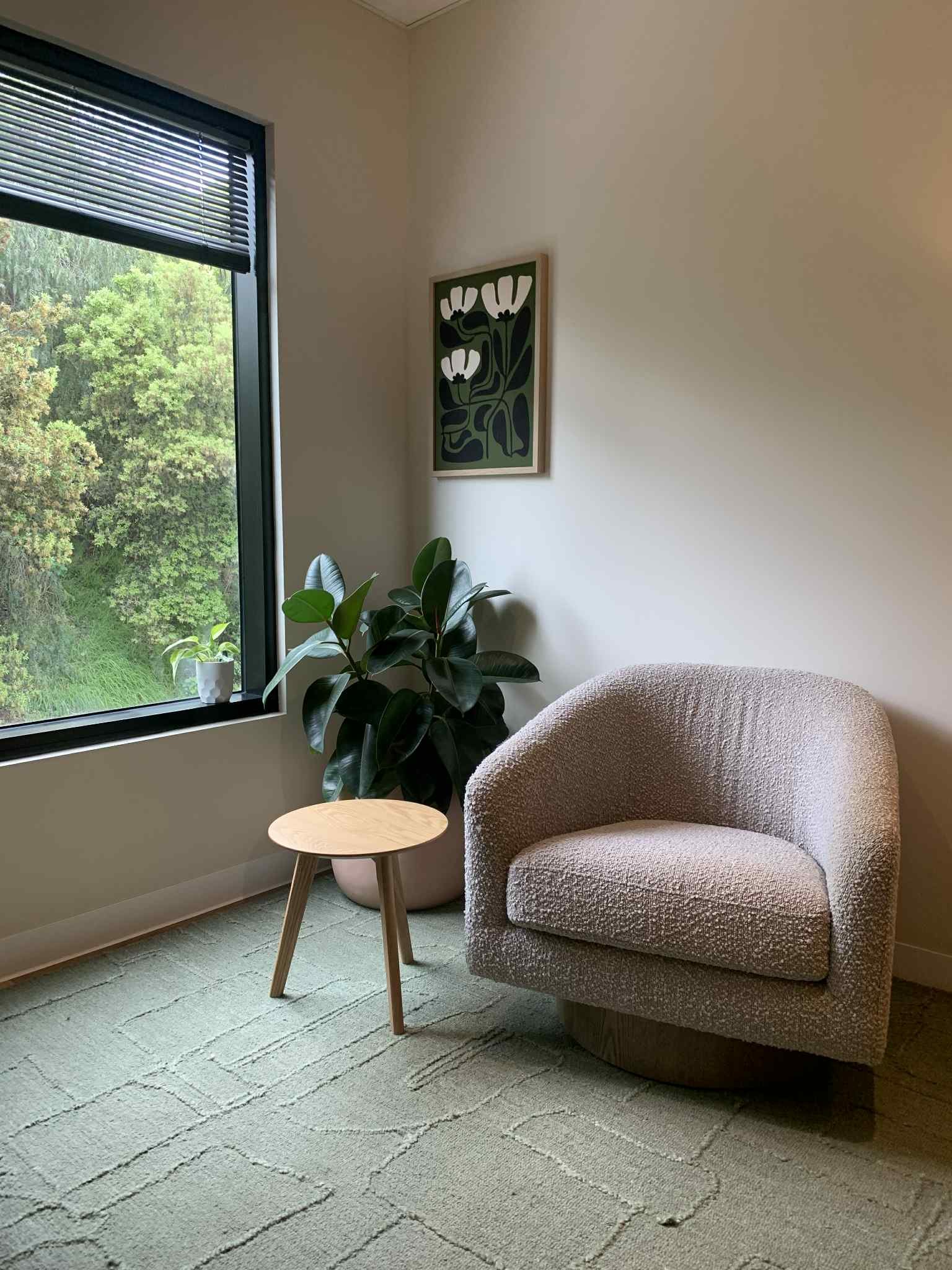 A cozy corner with a textured beige armchair, a small wooden round side table, a large leafy houseplant, a window with greenery outside, and a framed floral artwork on a white wall.