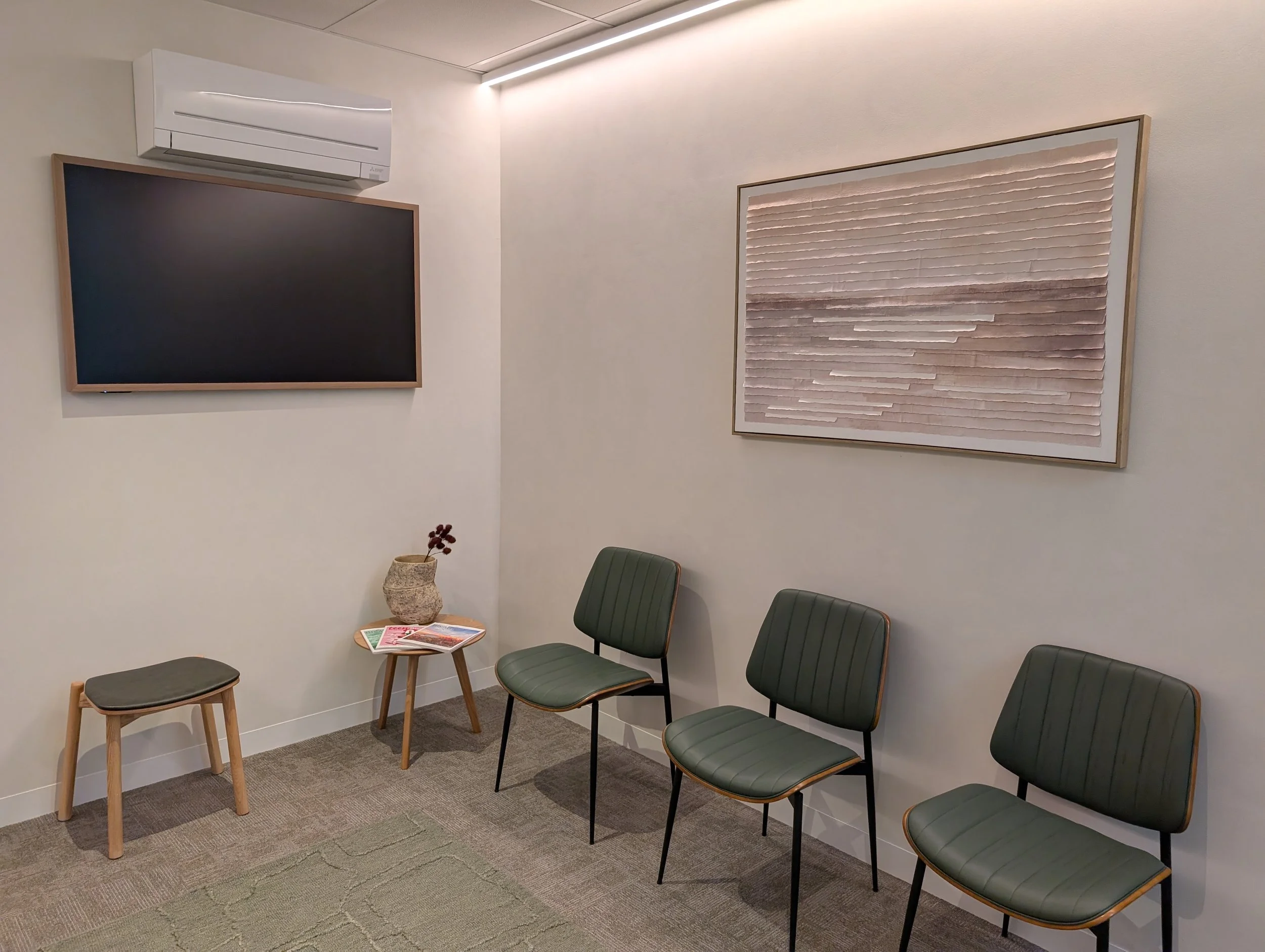 Waiting room with four green chairs, a small wooden table with magazines and a vase, a large framed artwork on the wall, a TV, and an air conditioning unit on the wall.