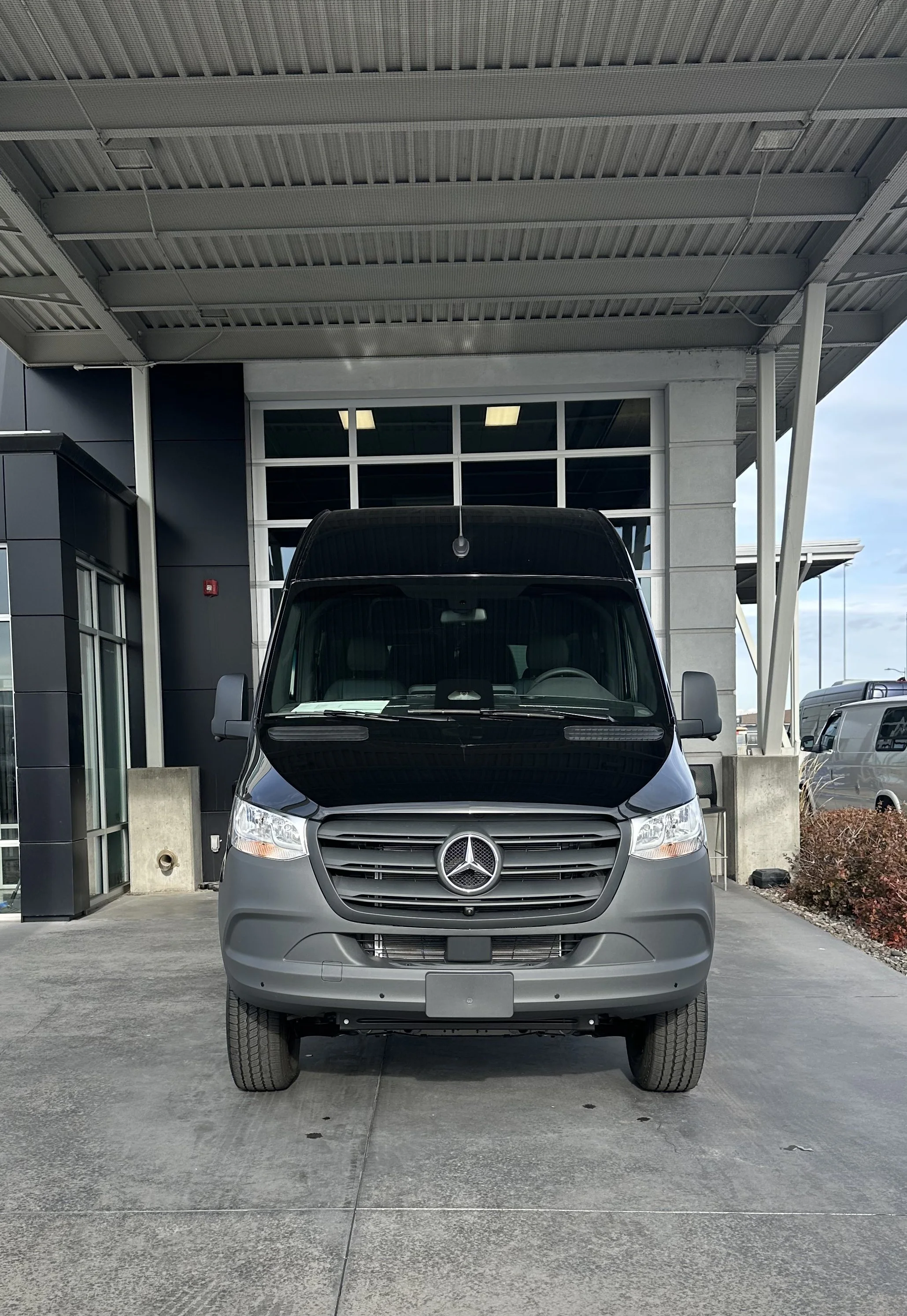 Front view of a gray Mercedes-Benz van parked outside a building with large glass windows and a metal roof.