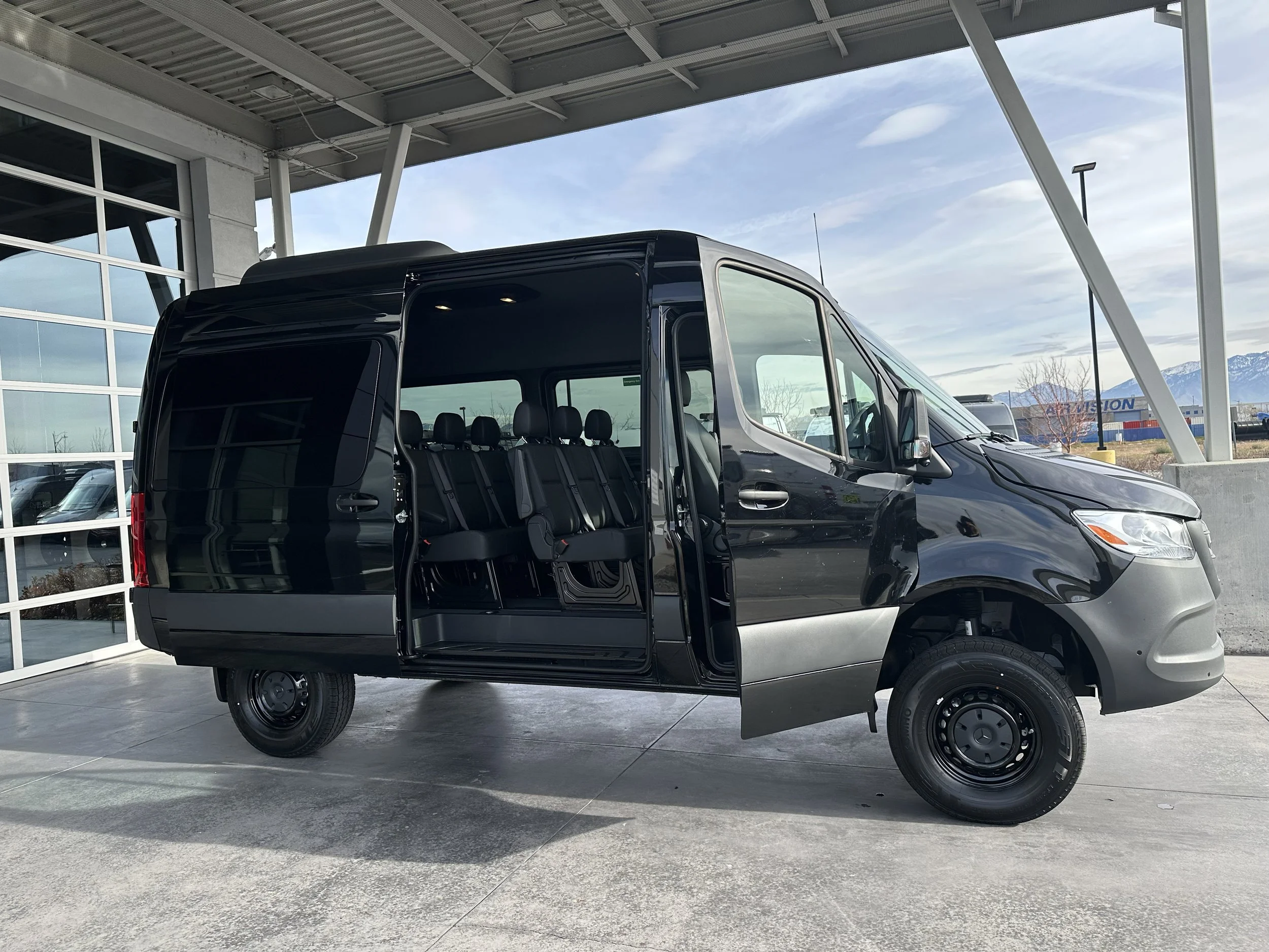 Black cargo van with side door open, showing interior seating, parked outside a building with glass windows and mountains in the distance.